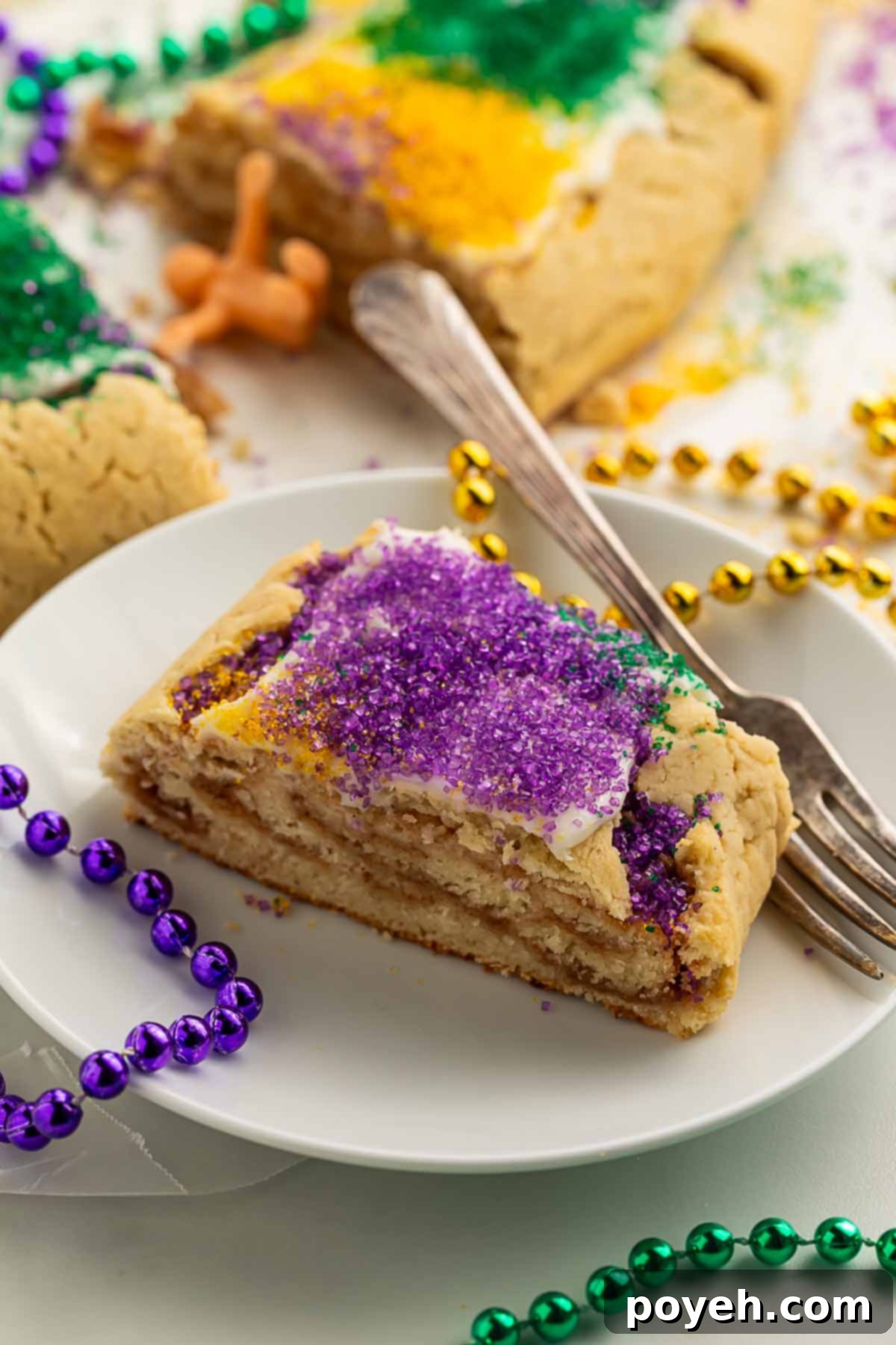 A piece of gluten free king cake, topped with rich purple sanding sugar, served on a white plate with a silver fork, surrounded by purple, gold, and green Mardi Gras bead necklaces. 