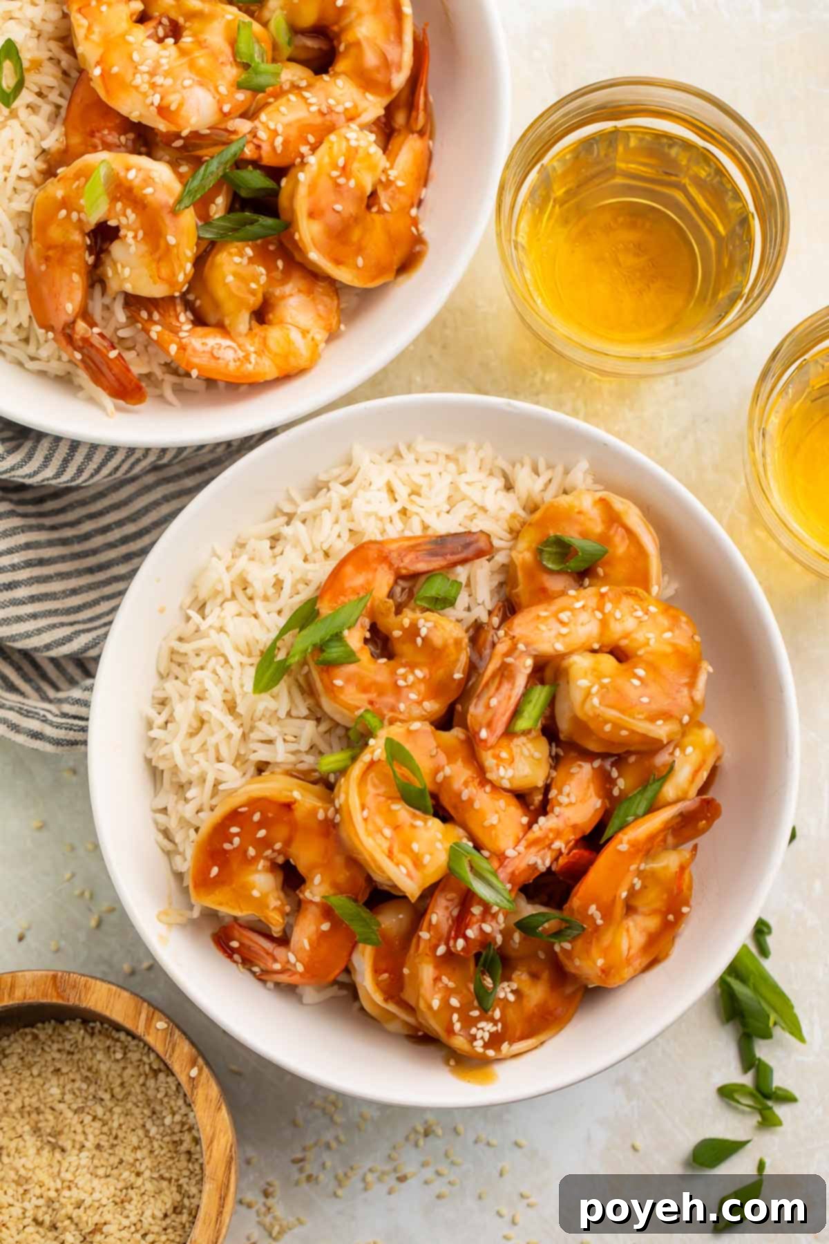 Bowls of teriyaki shrimp and cooked white rice on a table surrounded by green onions and a small bowl of sesame seeds, ready for a delicious meal.