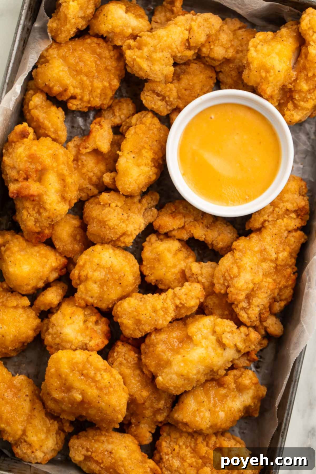 A pan lined with parchment paper holding air fryer cooked popcorn chicken and a ramekin of honey mustard sauce.
