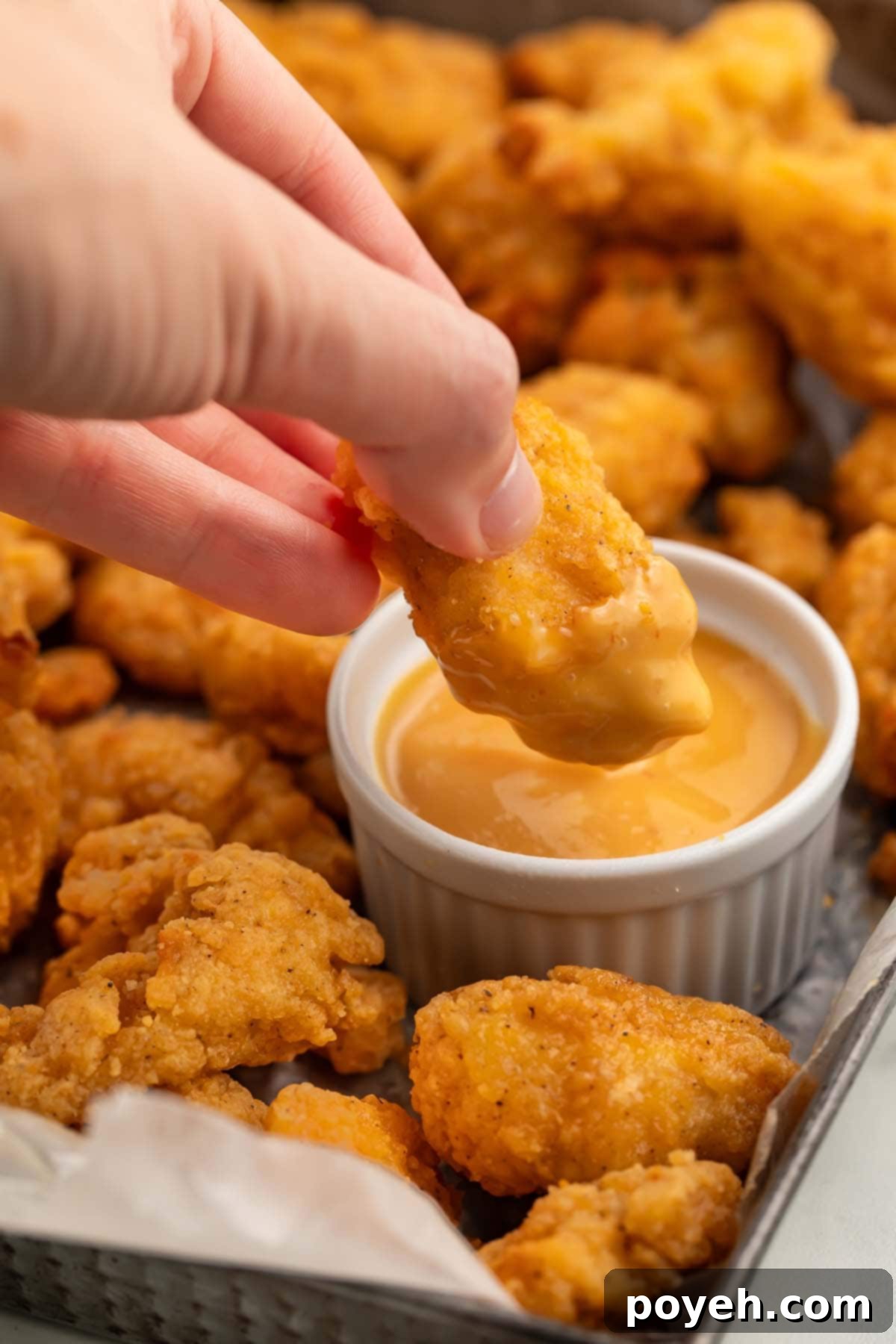 A white woman's hand holding a piece of air fryer popcorn chicken as she dips the chicken into a ramekin of honey mustard sauce.