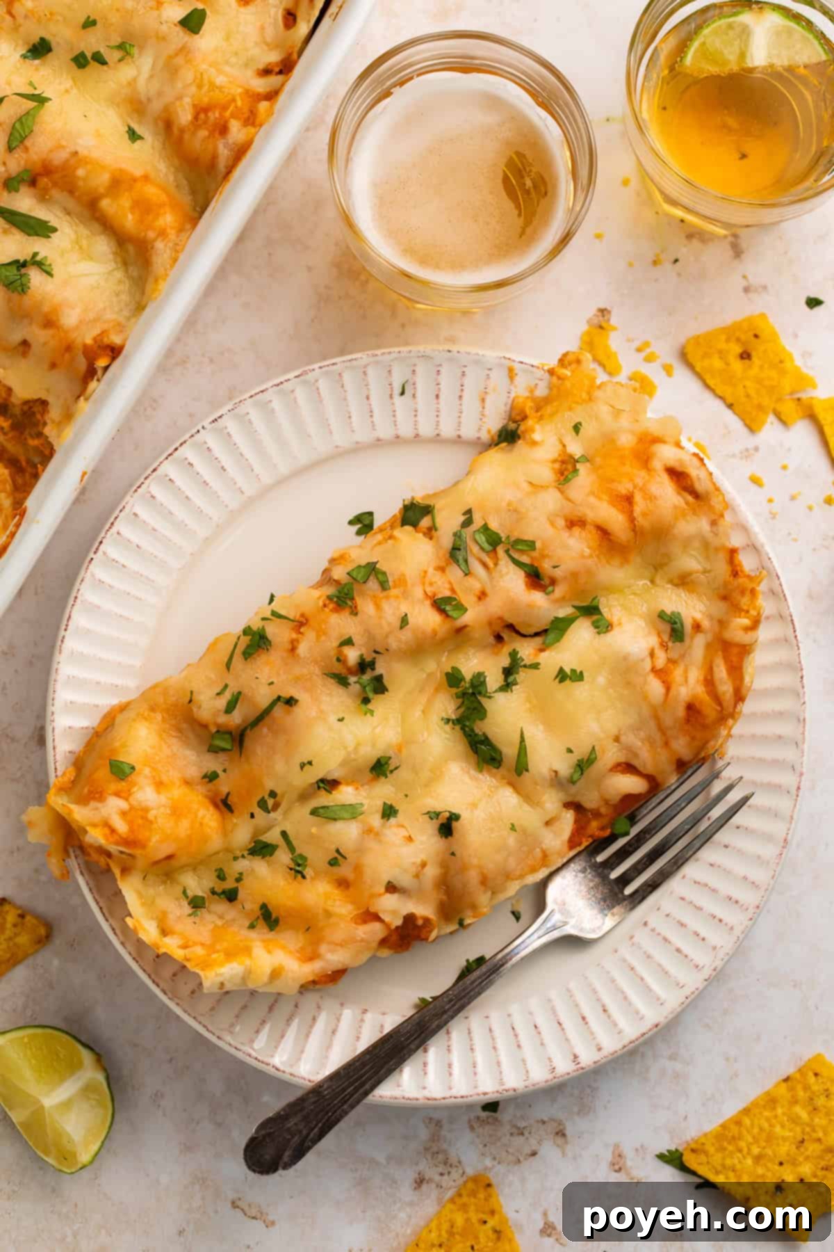 Two golden-brown creamy chicken enchiladas, generously topped with melted cheese, resting on a pristine white plate beside a fork. In the background, subtly blurred, are glasses of refreshing beverages and the baking dish filled with more enchiladas, suggesting a festive and inviting meal setting.