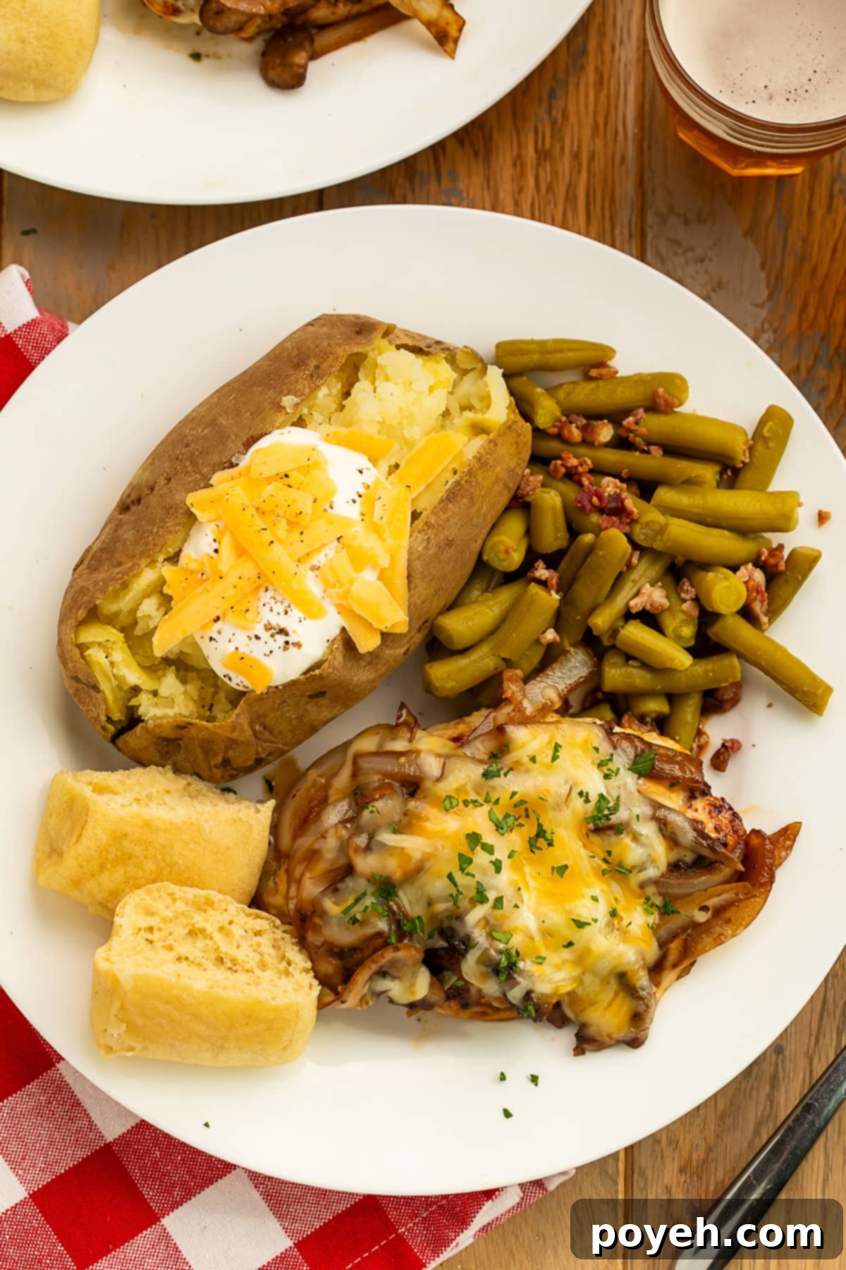 Plate of smothered chicken, green beans with bacon, a split baked potato topped with soured cream and grated cheese, and a torn bread roll.