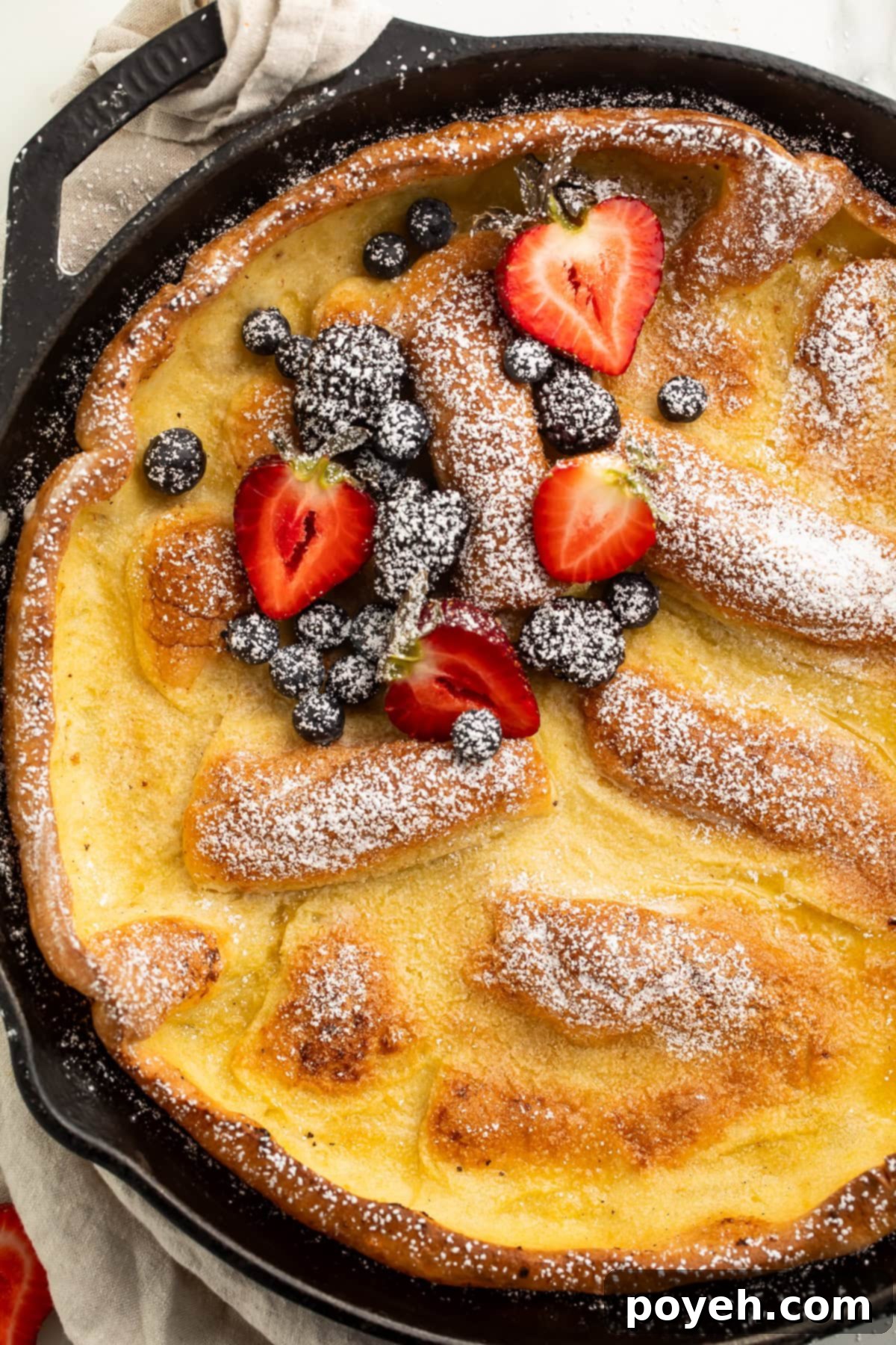 Close-up view of a perfectly puffed Gluten-Free Dutch Baby Pancake adorned with vibrant strawberries and blueberries, nestled in its cast iron skillet.
