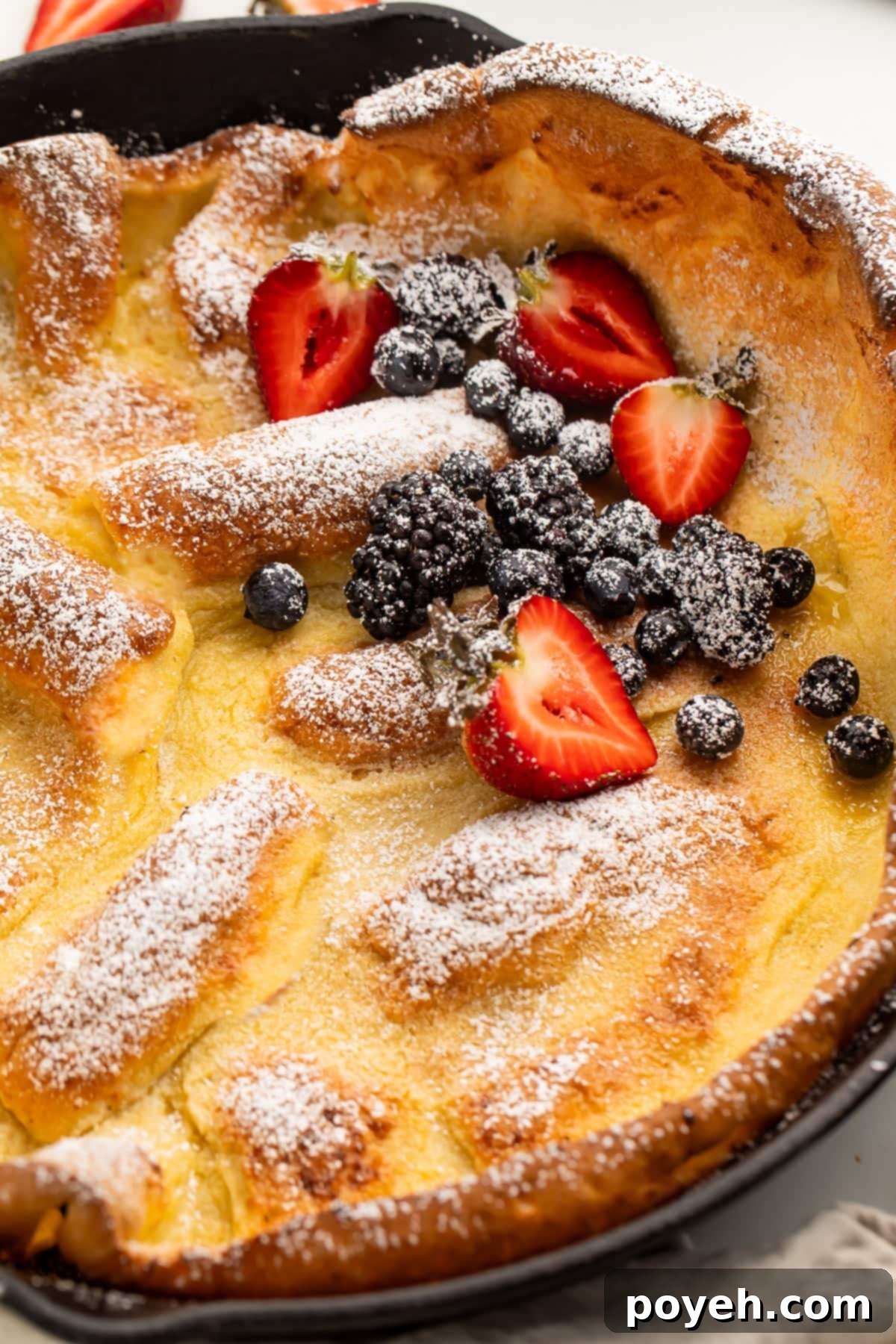 A beautifully pillowy Gluten-Free Dutch Baby Pancake, generously finished with a dusting of powdered sugar and a colorful medley of fresh berries, ready to be served.
