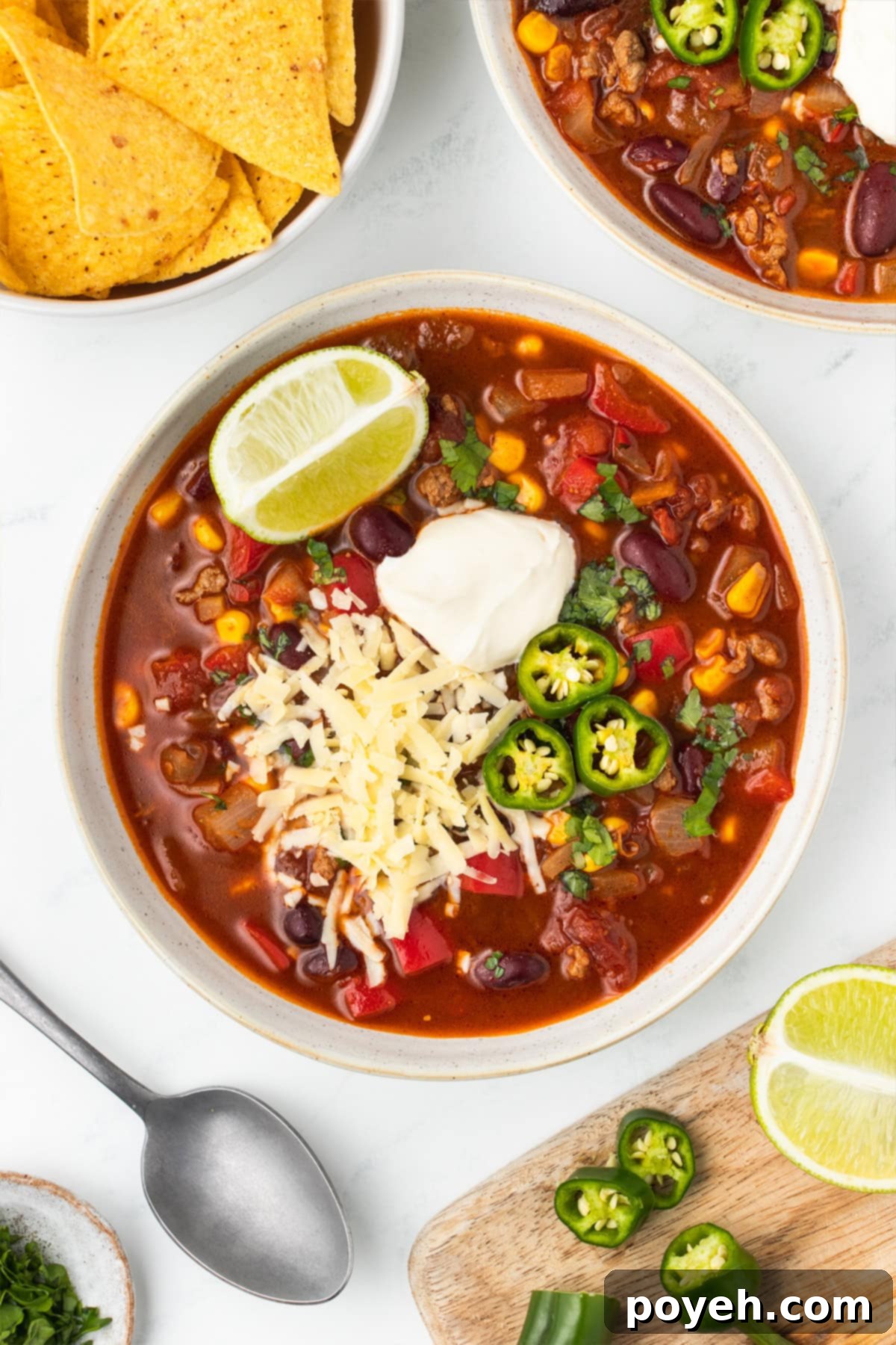 A vibrant bowl of chili with corn, garnished with a dollop of sour cream, shredded cheese, thinly sliced jalapeños, and a fresh lime wedge.