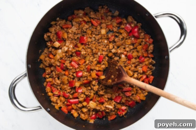 Ground beef browning in a pot with sautéed onions and peppers, being broken up with a wooden spoon.