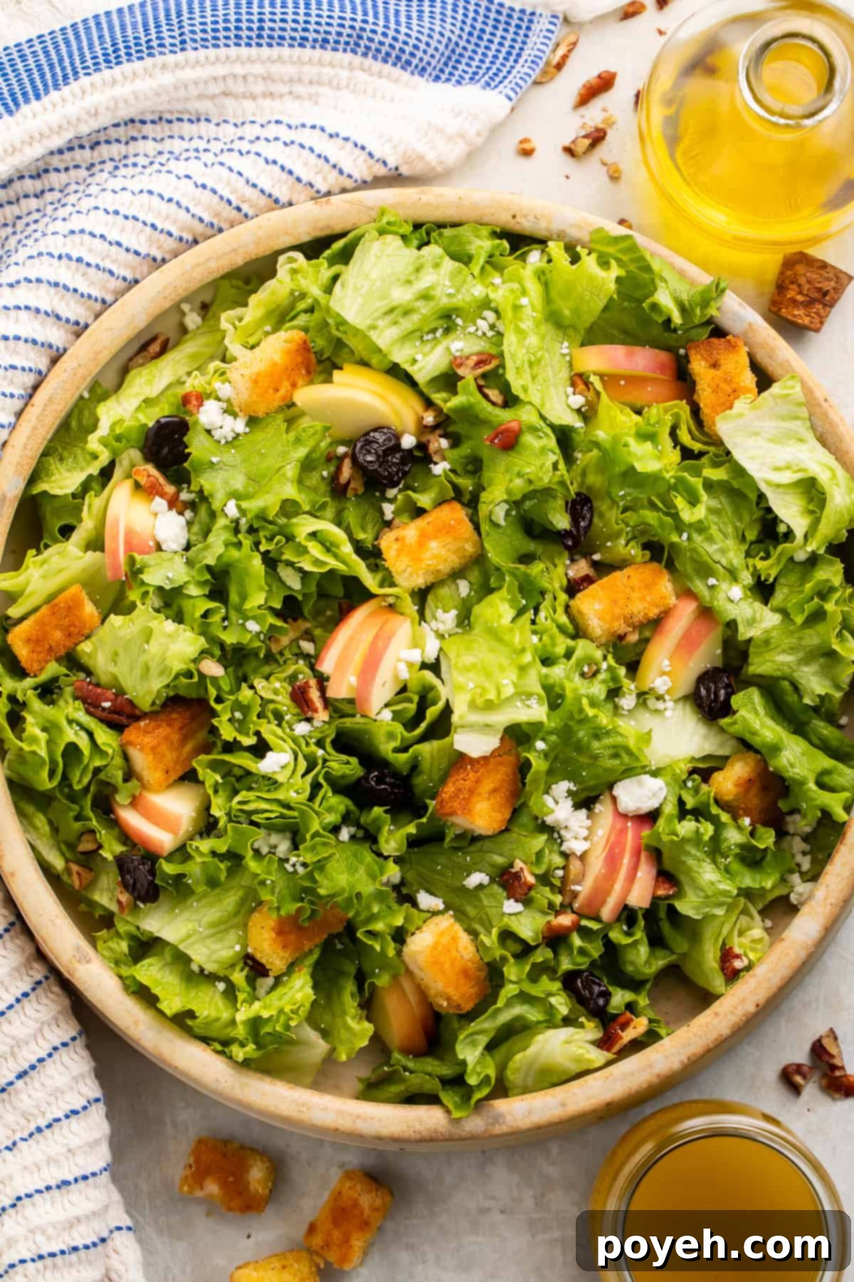 Wooden bowl of fresh green salad topped with sliced apples and golden gluten-free croutons, with a jug of yellow dressing on the side.