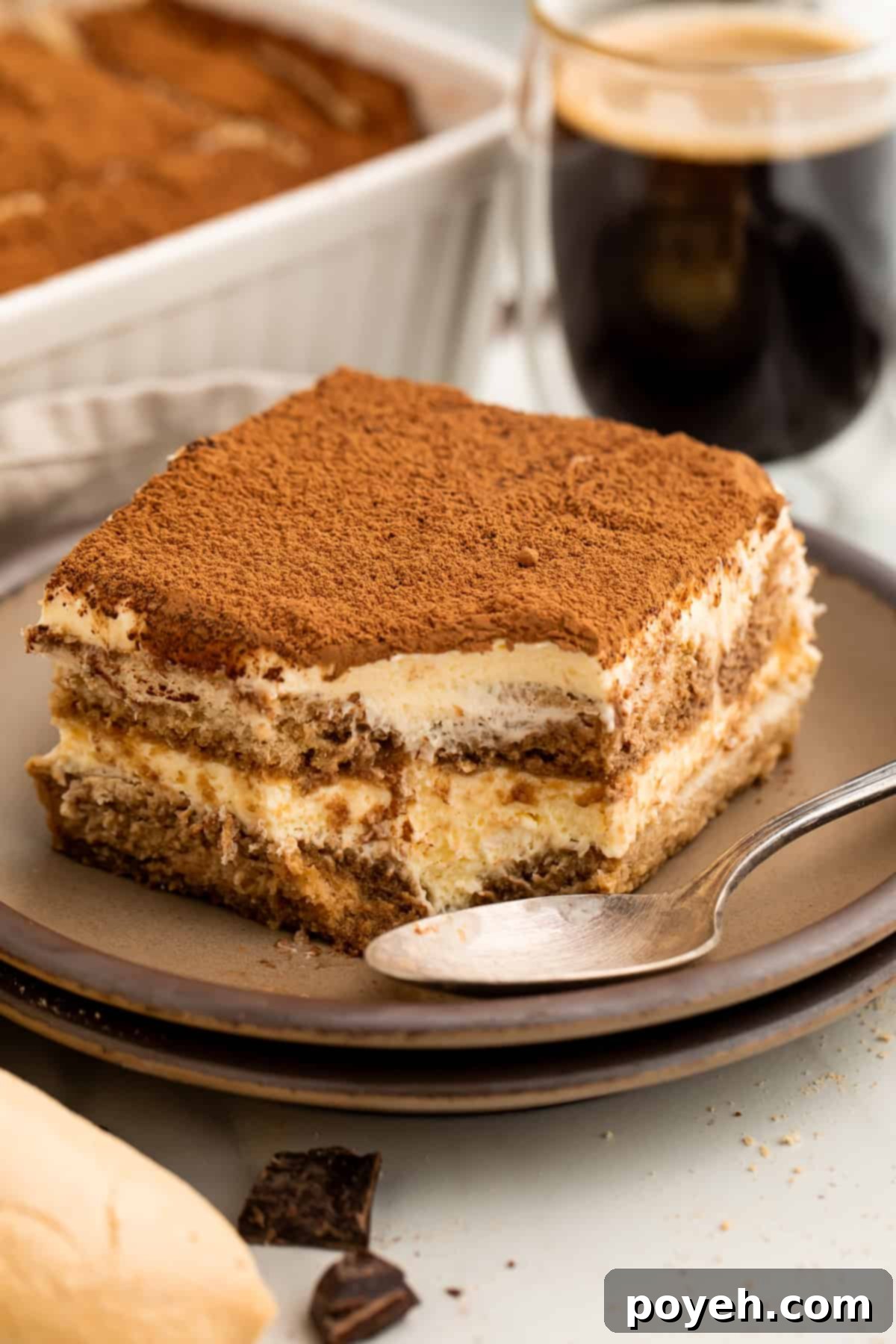 Slice of tiramisu on a plate with a spoon where a spoonful has just been taken out of the corner.