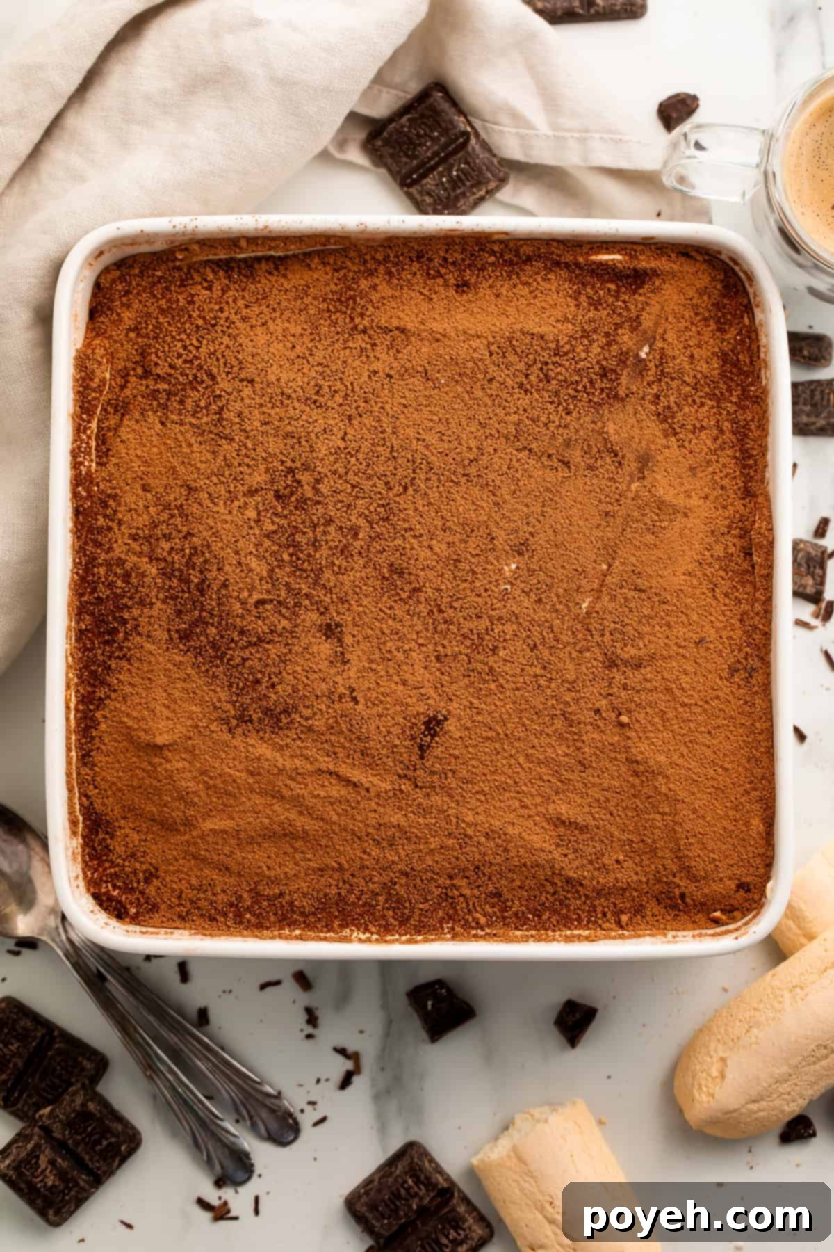 Overhead view of a gluten free tiramisu in a white baking dish with biscuits and dark chocolate squares scattered around. 