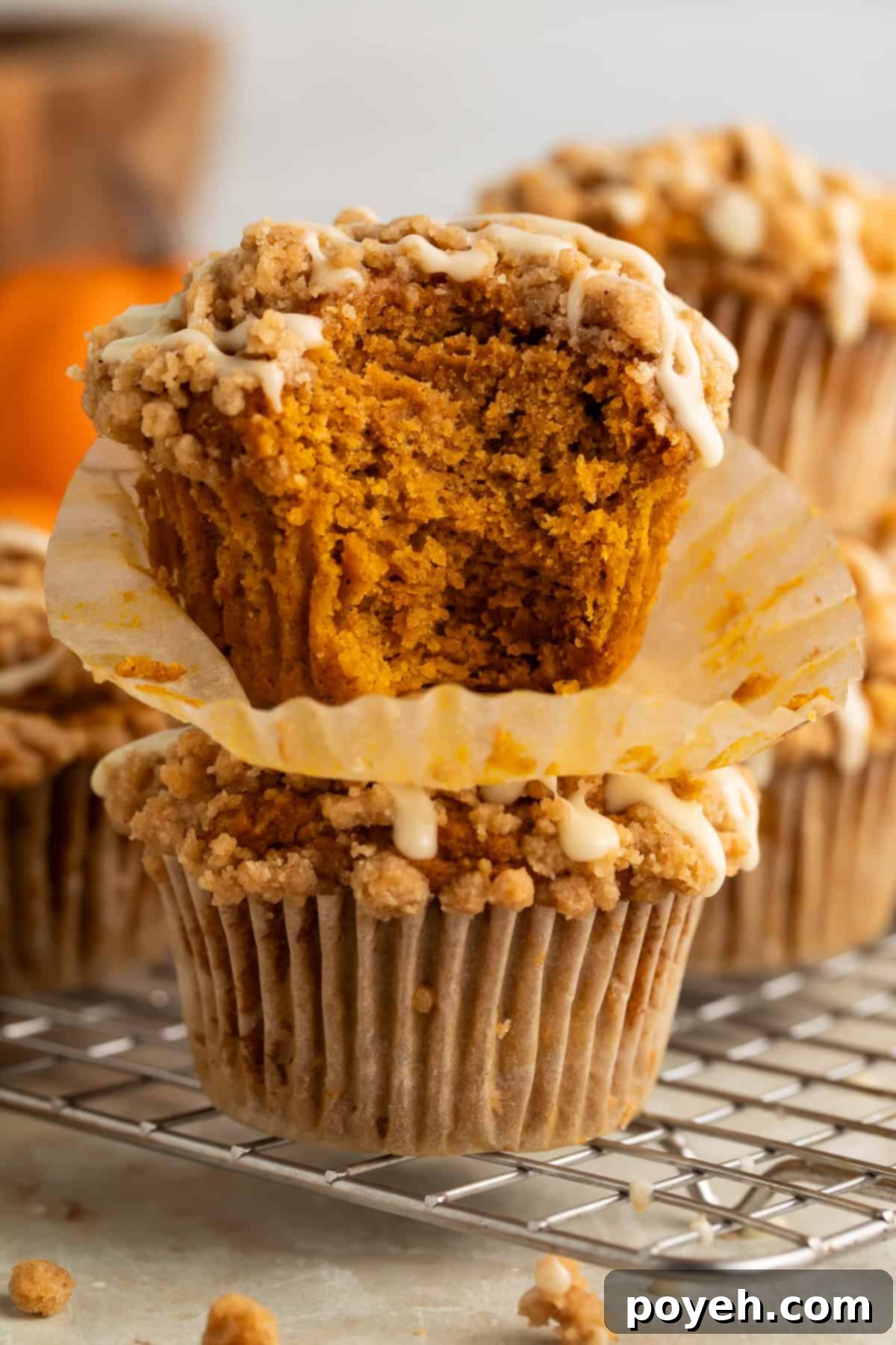 Two golden-brown gluten-free pumpkin muffins, one stacked on top of the other. A bite has been taken from the upper muffin, revealing its moist, spiced interior and hinting at the crunchy streusel and glossy maple glaze.