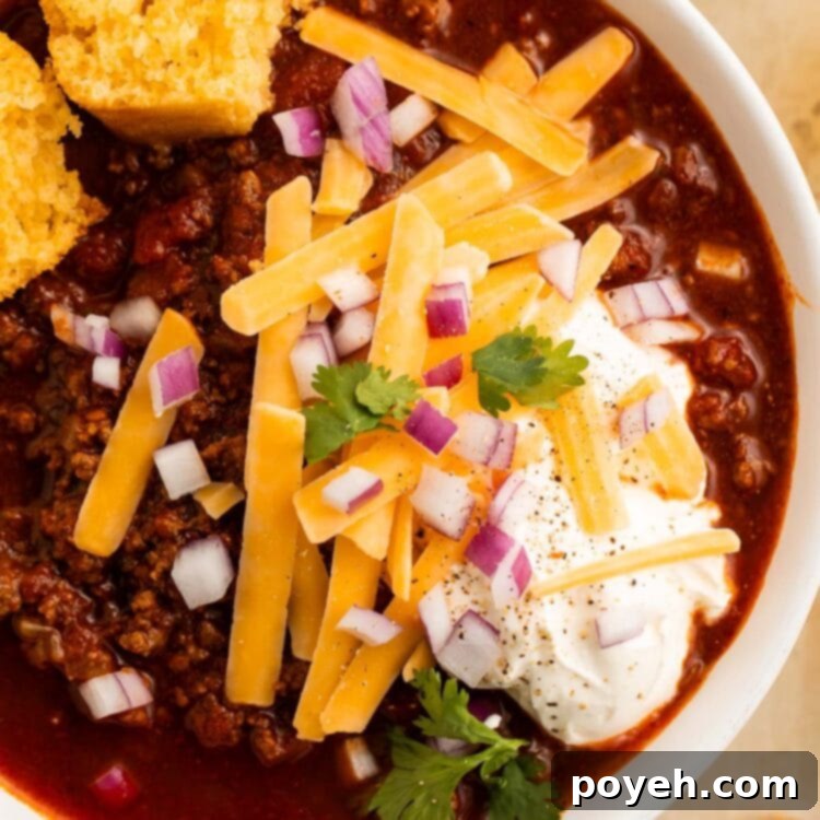 Close-up of a white bowl containing deep red low carb chili topped with a large dollop of sour cream, thick shreds of yellow cheddar cheese, diced purple onion, and chopped parsley.