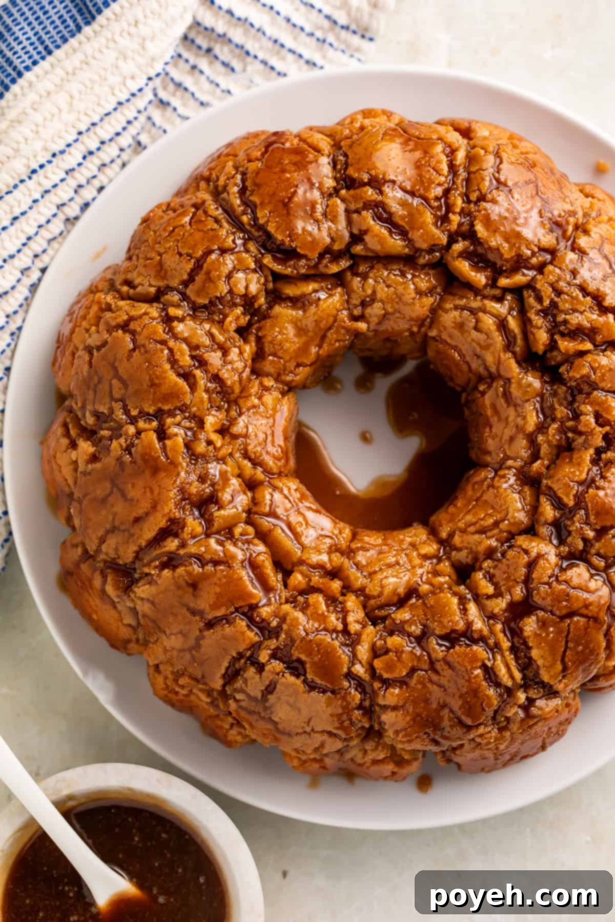 Gooey Gluten-Free Monkey Bread Bliss 3 Overhead view of a monkey bread drizzled with caramel on a white plate. A white bowl of caramel with a white spoon sits off to one side.