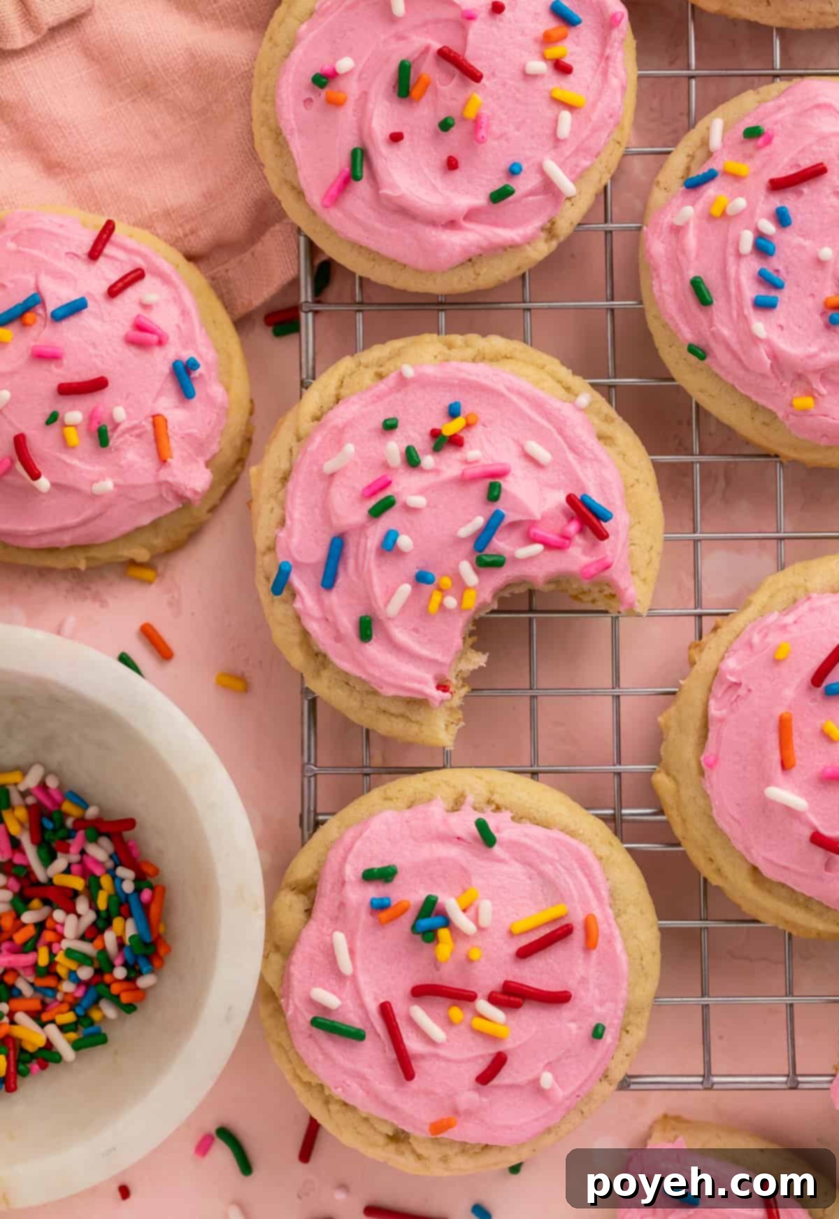 Pink frosted sugar cookies with rainbow sprinkles next to a bowl of sprinkles, one with a bite taken out.