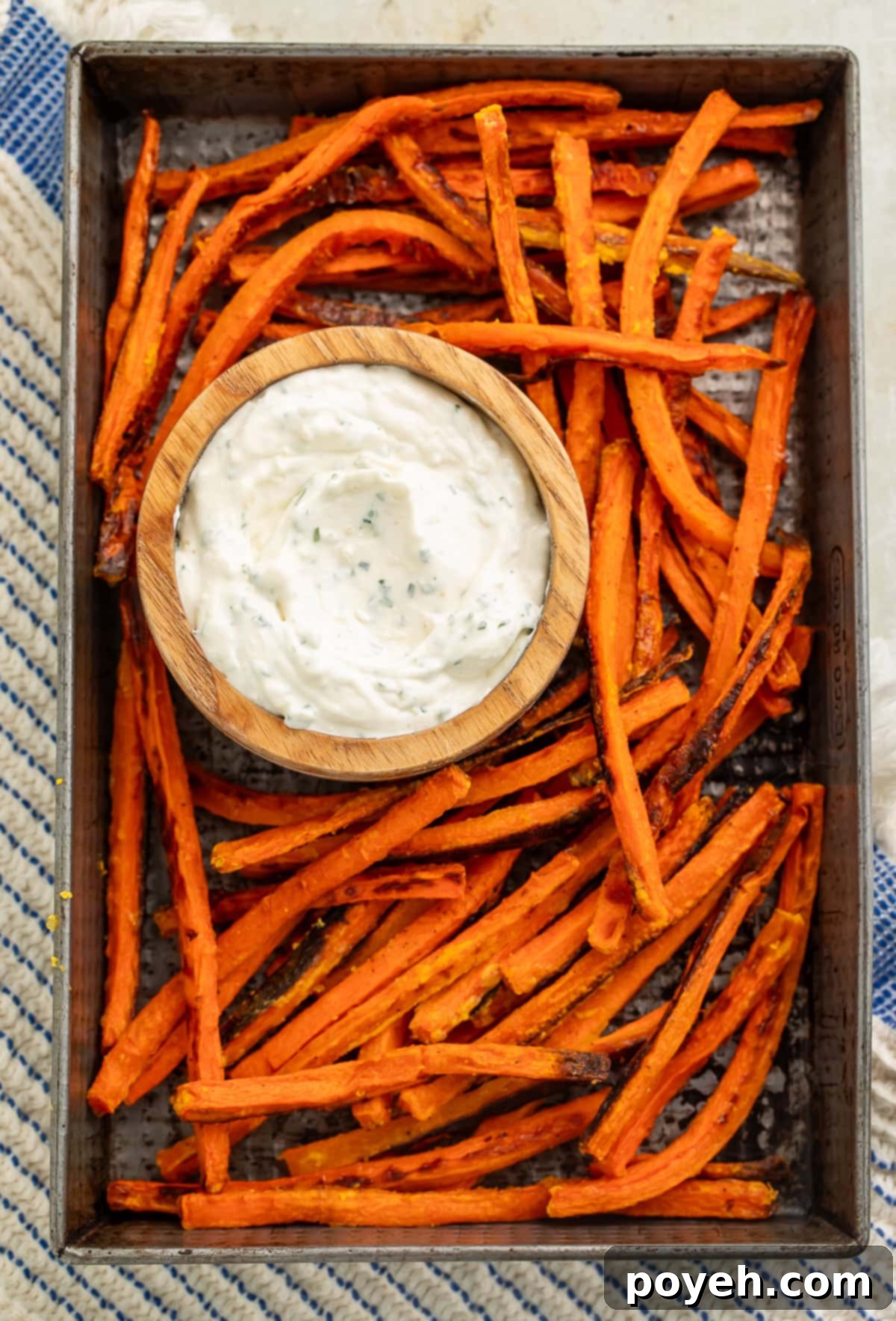 Top-down shot of a baking tray featuring an abundant serving of oven-baked carrot fries, with a rustic wooden bowl of creamy herb dip centered among them.