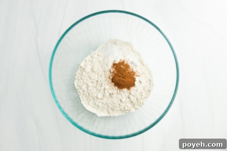 Overhead view of gluten-free flour, salt, cinnamon, and baking soda neatly arranged in a large glass mixing bowl.