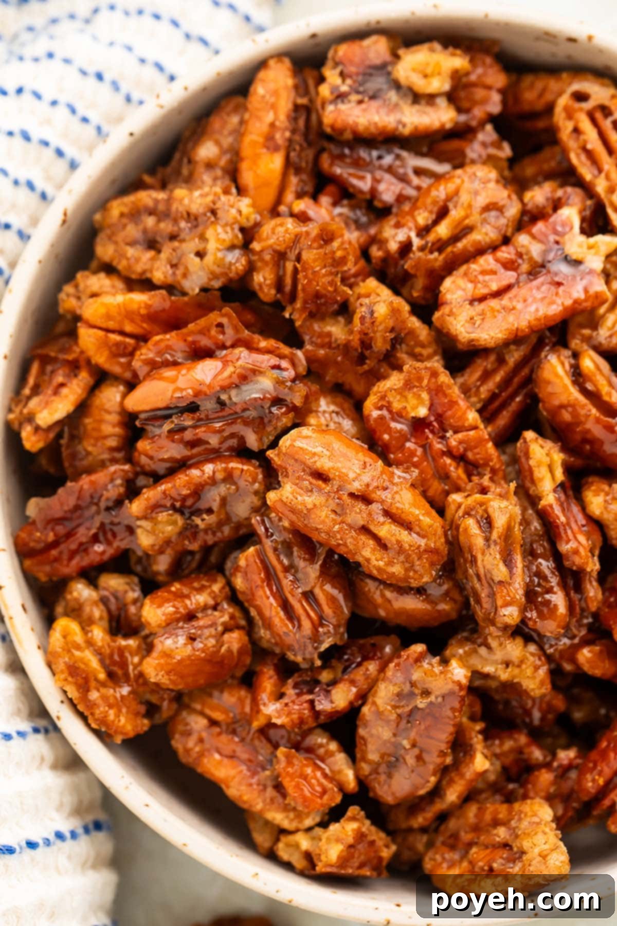 A bowl of maple-cinnamon glazed pecans sits on a table surrounded by a white and blue striped kitchen towel.
