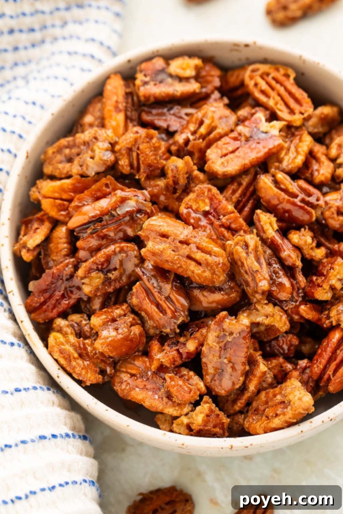 A bowl of maple-cinnamon glazed pecans sits on a table surrounded by a white and blue striped kitchen towel.