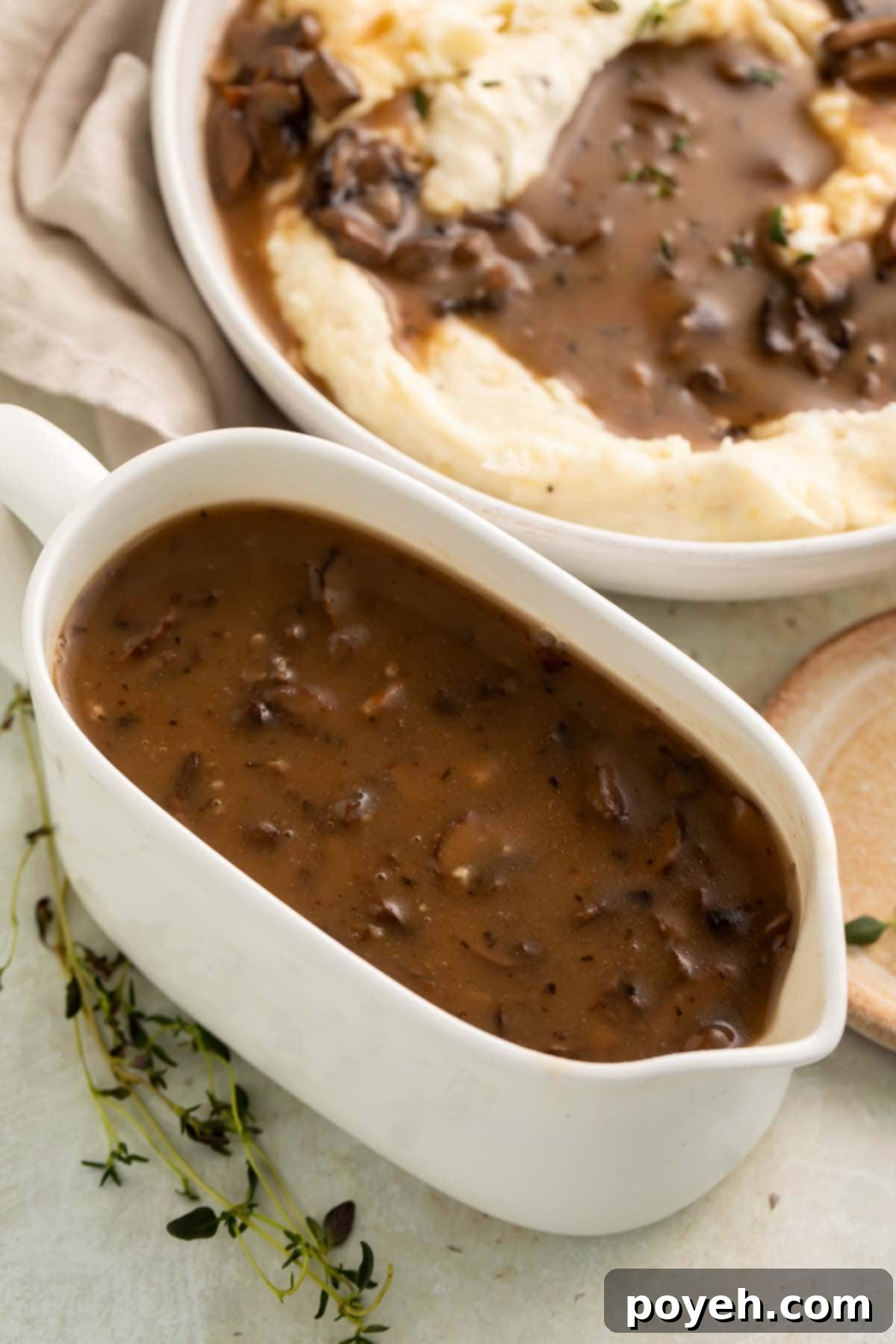 A elegant ceramic white gravy boat filled with rich, glossy vegan mushroom gravy, positioned next to a steaming bowl of fluffy mashed potatoes generously topped with the same savory gravy.