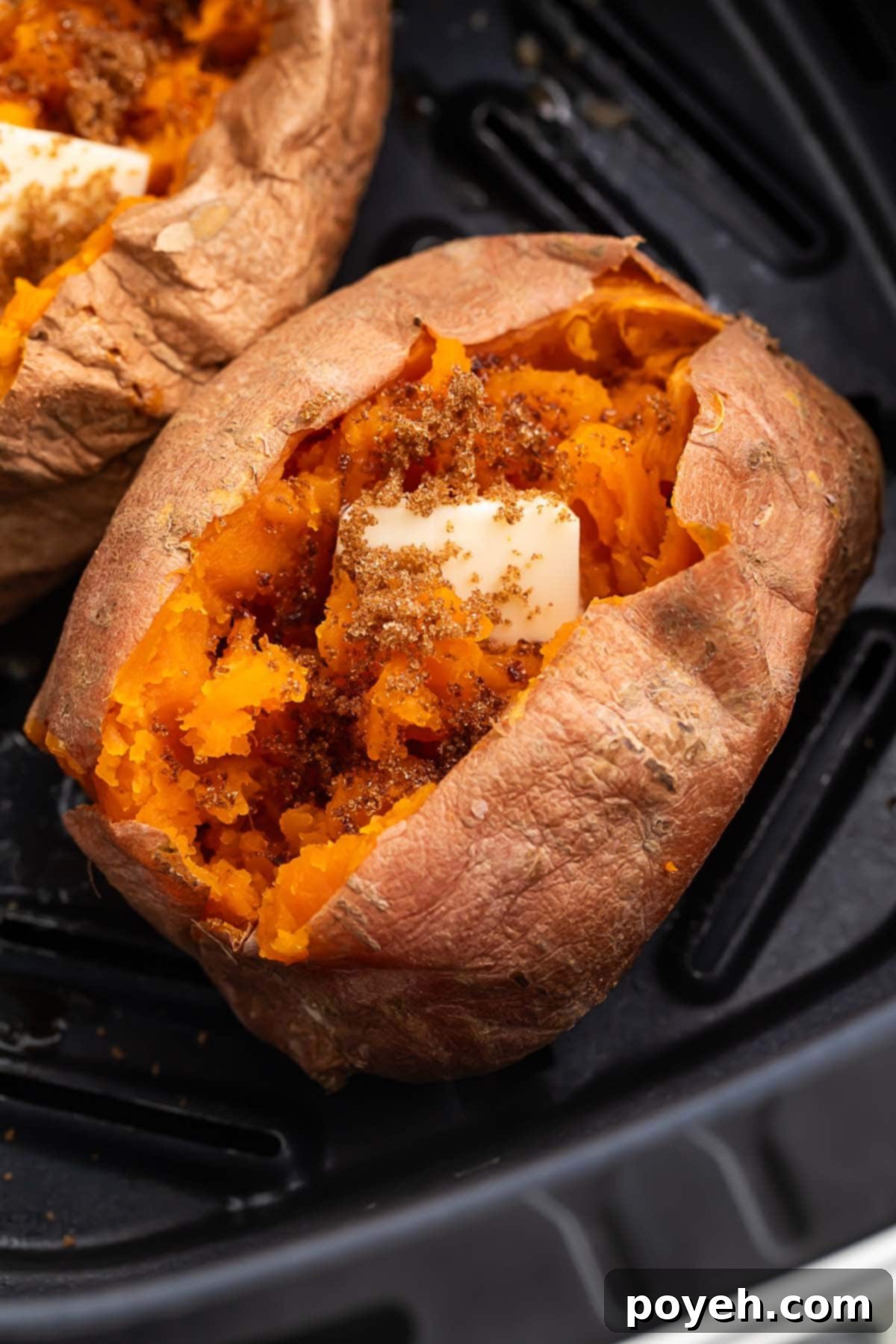 A cooked sweet potato resting in an air fryer basket. The top of the potato is split open, with the insides fluffed and topped with a pat of butter.