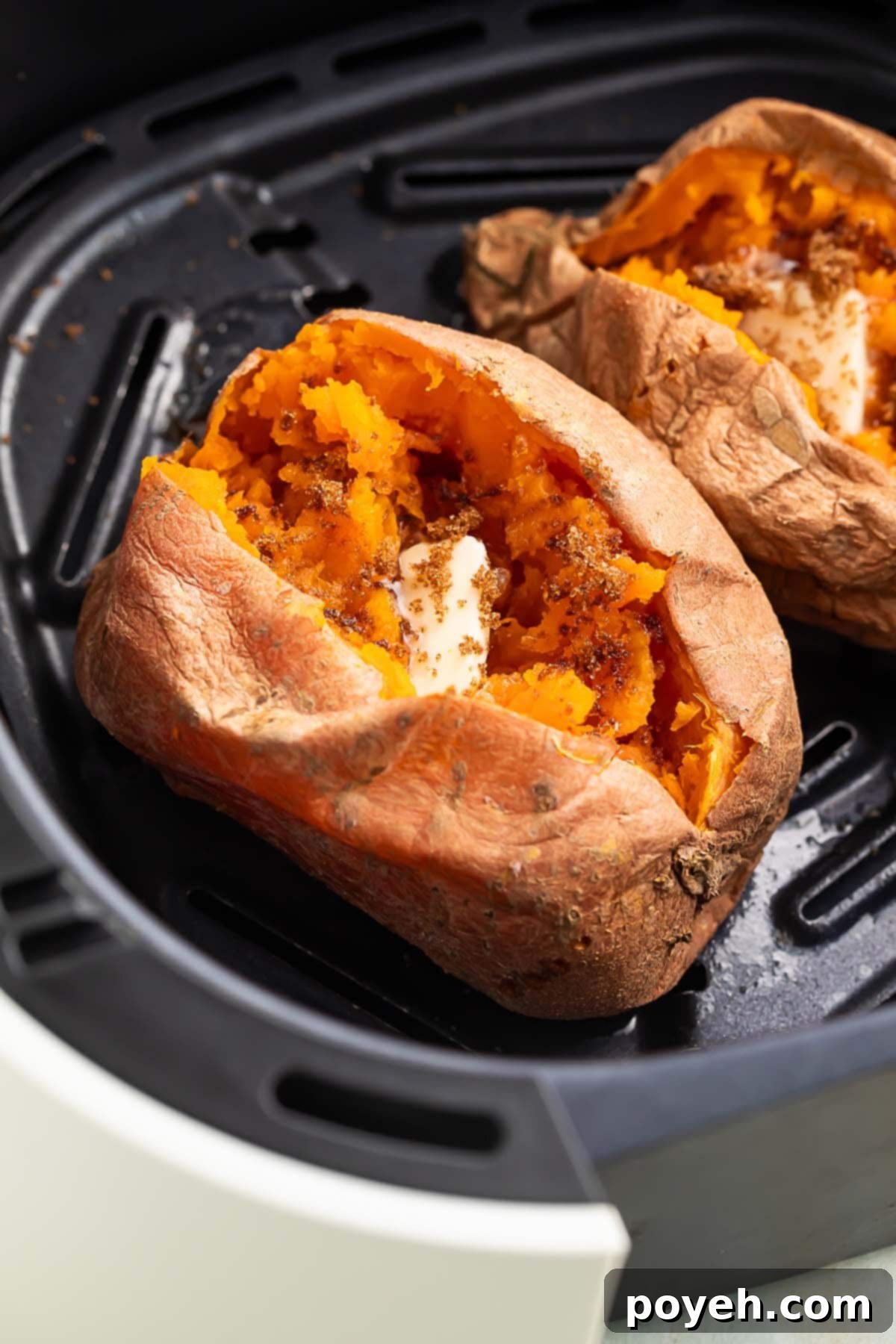 A cooked sweet potato resting in an air fryer basket. The top of the potato is split open, with the insides fluffed and topped with a pat of butter.