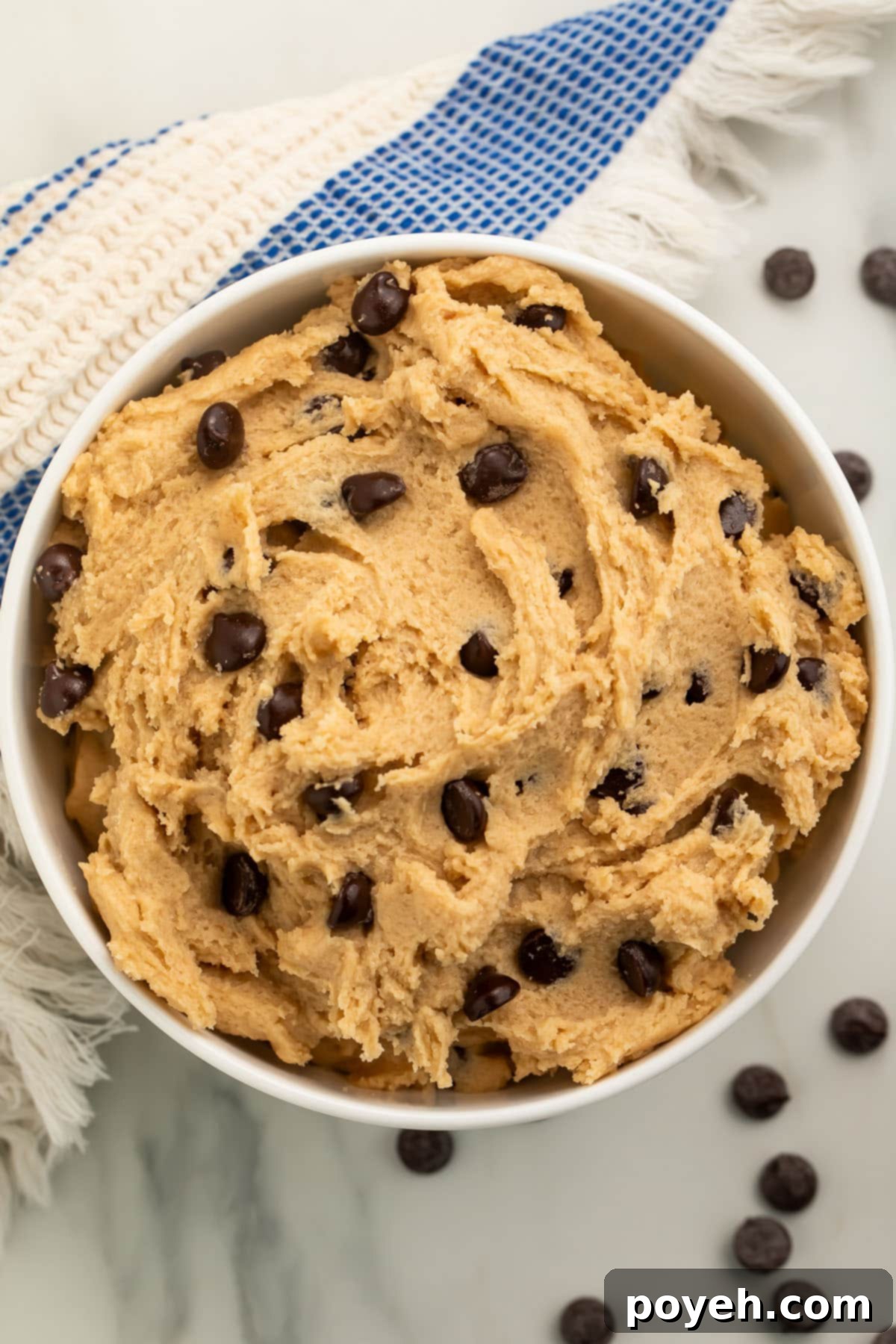 A beautifully styled scene featuring a bowl of vegan cookie dough on a kitchen counter, surrounded by loose chocolate chips and a charming blue and white striped kitchen towel.