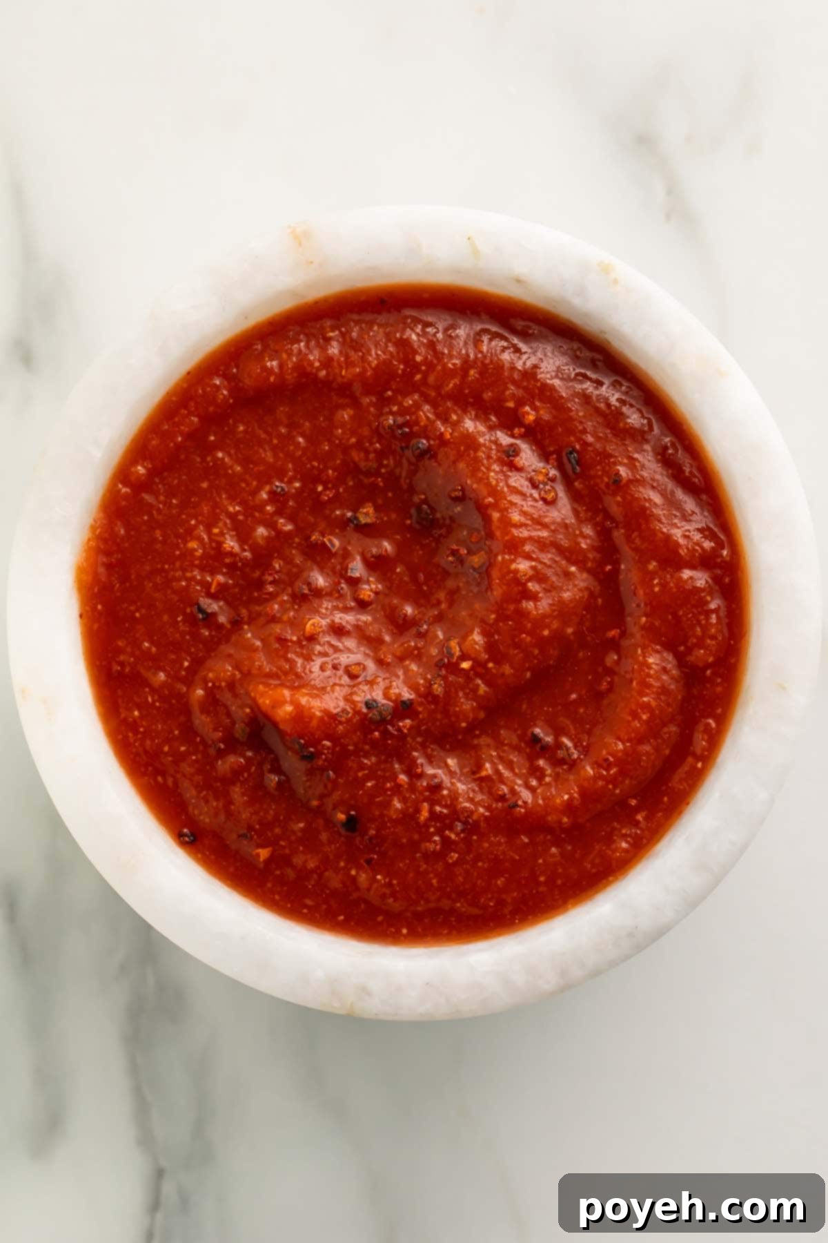 Top-down view of a small white bowl of deep red taco sauce on a marble countertop.