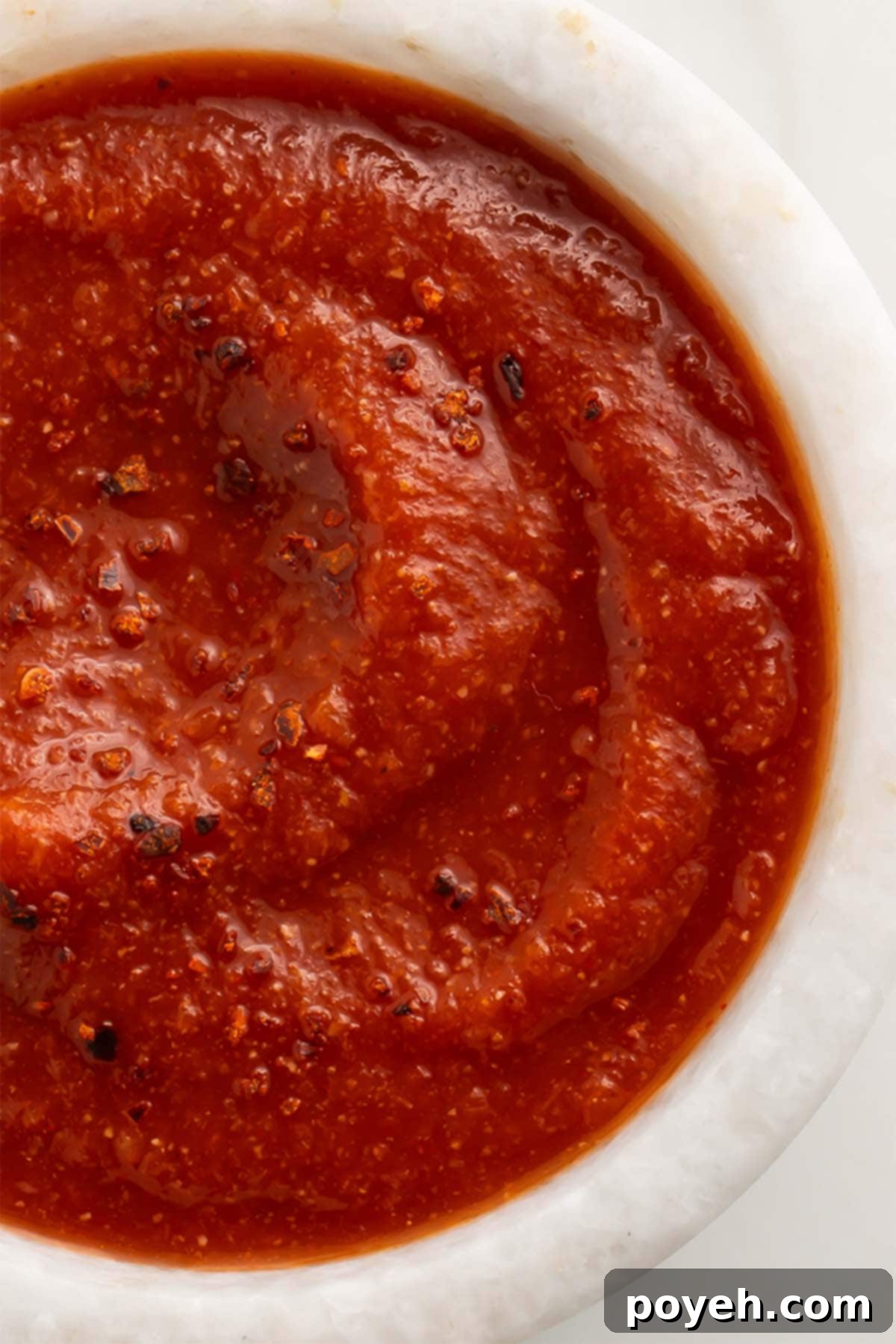 Top-down view of a small white bowl of deep red taco sauce on a marble countertop.