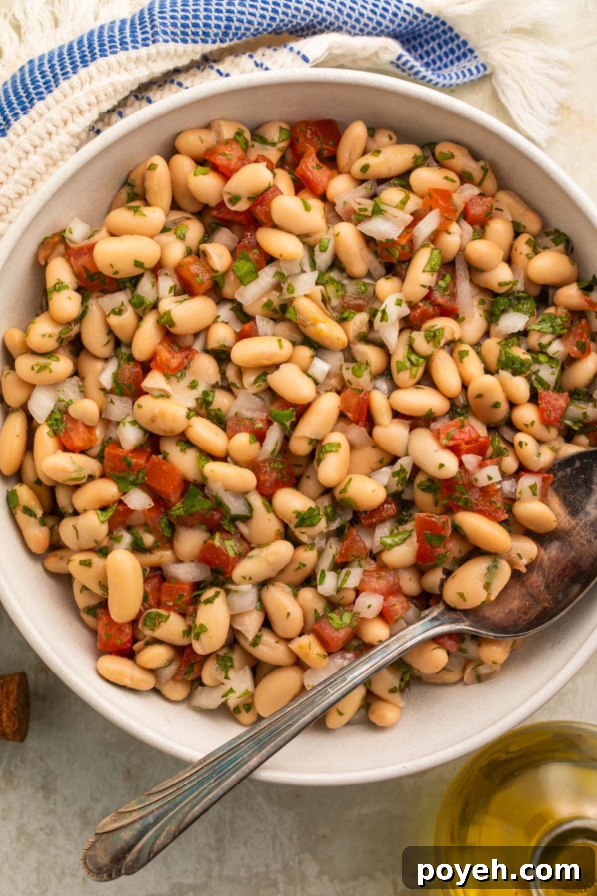 Mediterranean White Bean Salad 3 A large white serving bowl overflowing with a delightful white bean salad, featuring a generous mix of tender white beans, vibrant red tomatoes, thinly sliced red onions, and freshly chopped green parsley, all lightly dressed.