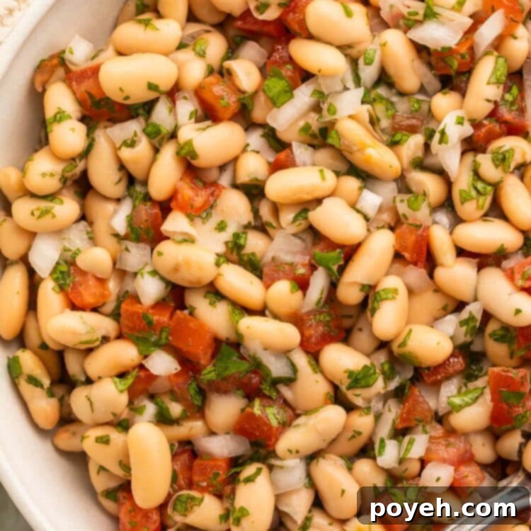 Mediterranean White Bean Salad 5 A beautifully presented large white bowl of white bean salad, featuring a harmonious blend of onions, tomatoes, fresh parsley, and tender white beans, all coated in a light, glossy vinaigrette, ready for serving.