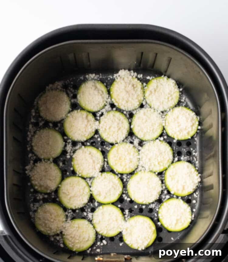 Zucchini chips generously topped with grated Parmesan cheese inside the air fryer basket before cooking.