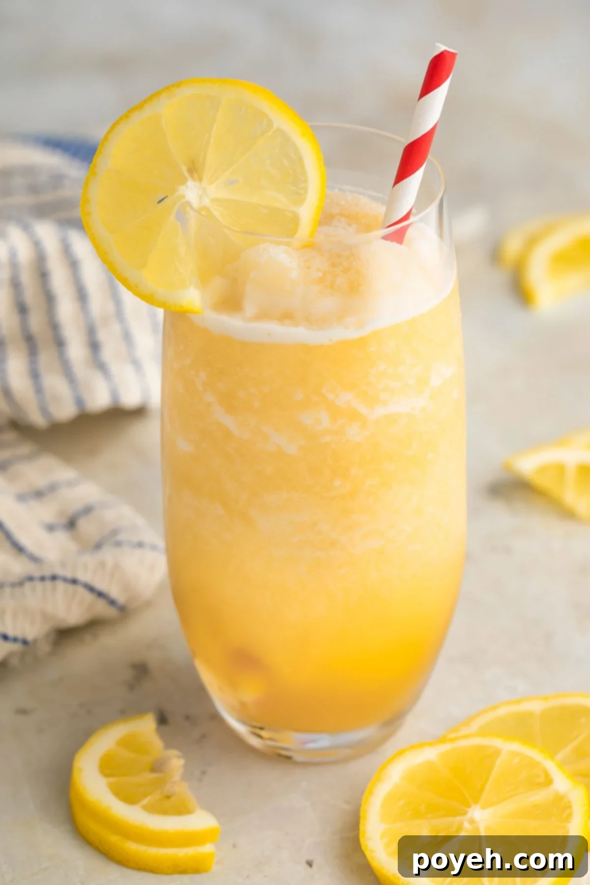 A tall glass filled with vibrant yellow frozen lemonade, elegantly garnished with a fresh lemon slice and a classic red & white striped straw. Perfect for a refreshing summer day.