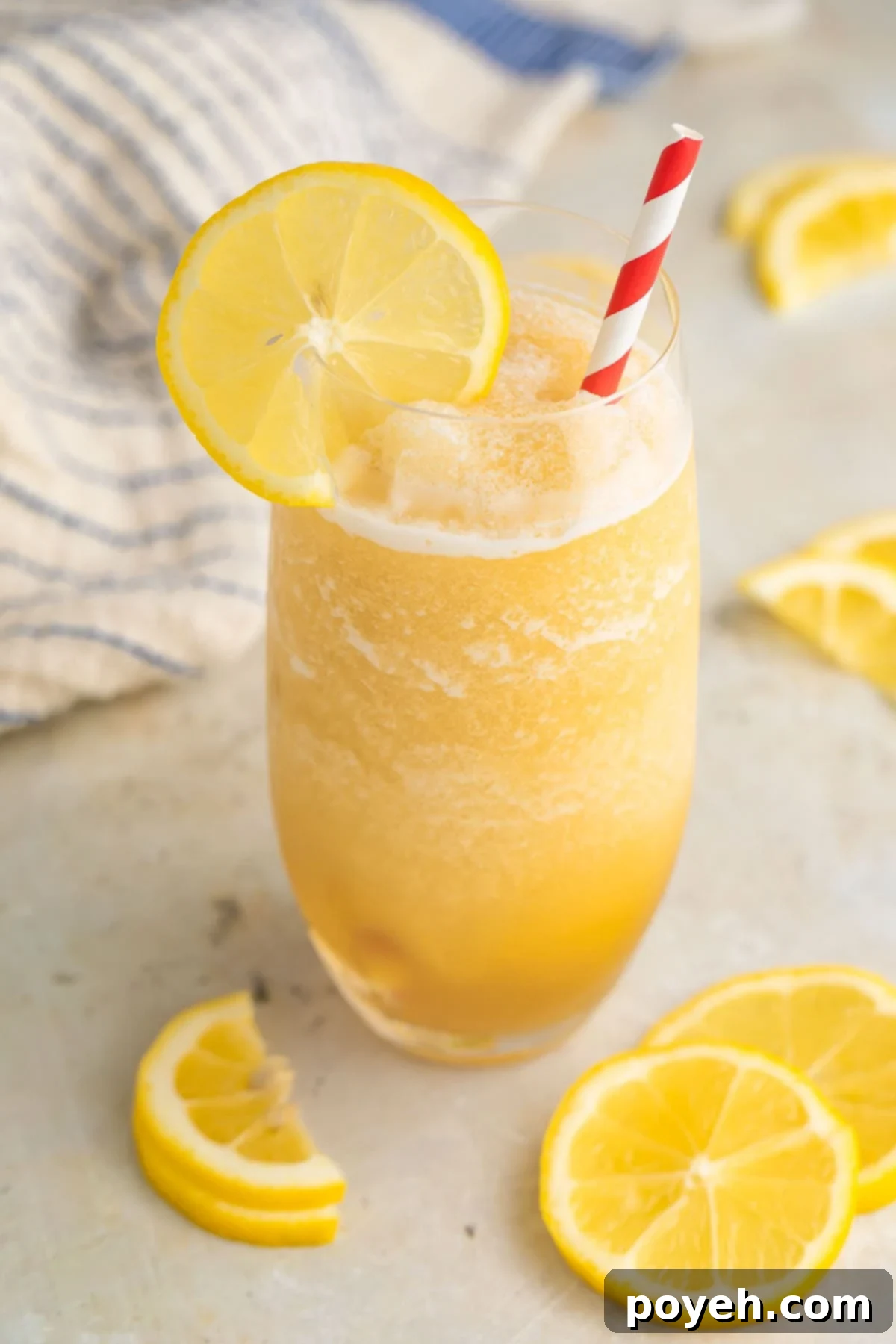 A tall glass brimming with golden frozen lemonade, beautifully garnished with a vibrant lemon wheel and a festive red & white striped straw, inviting you to take a refreshing sip.