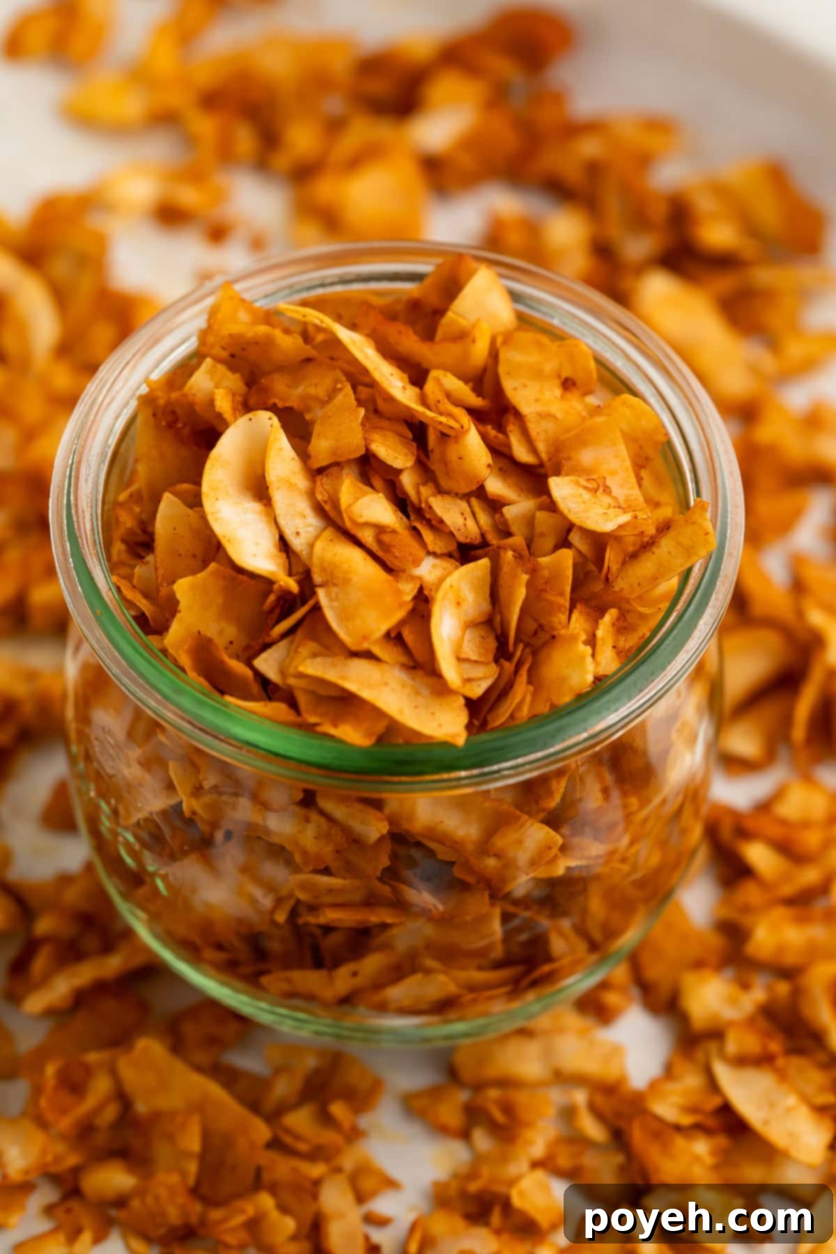 A beautifully styled glass jar filled with golden-brown, crispy coconut bacon chips, surrounded by loose coconut bacon pieces on a rustic table surface.