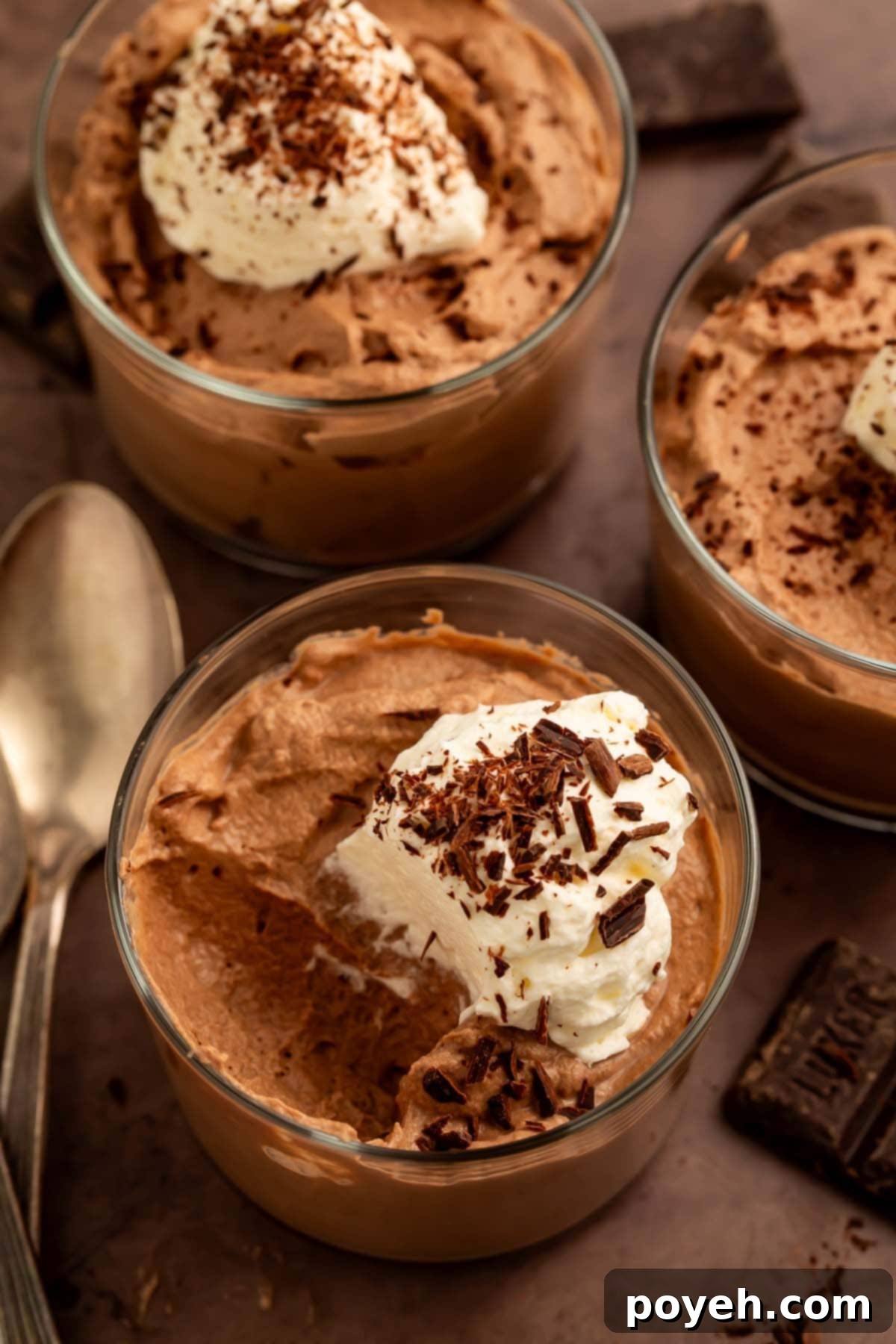 Close-up shot of three glass cups of keto chocolate mousse, exquisitely garnished with a generous dollop of sugar-free whipped cream and delicate chocolate shavings, highlighting its rich texture and enticing appeal.