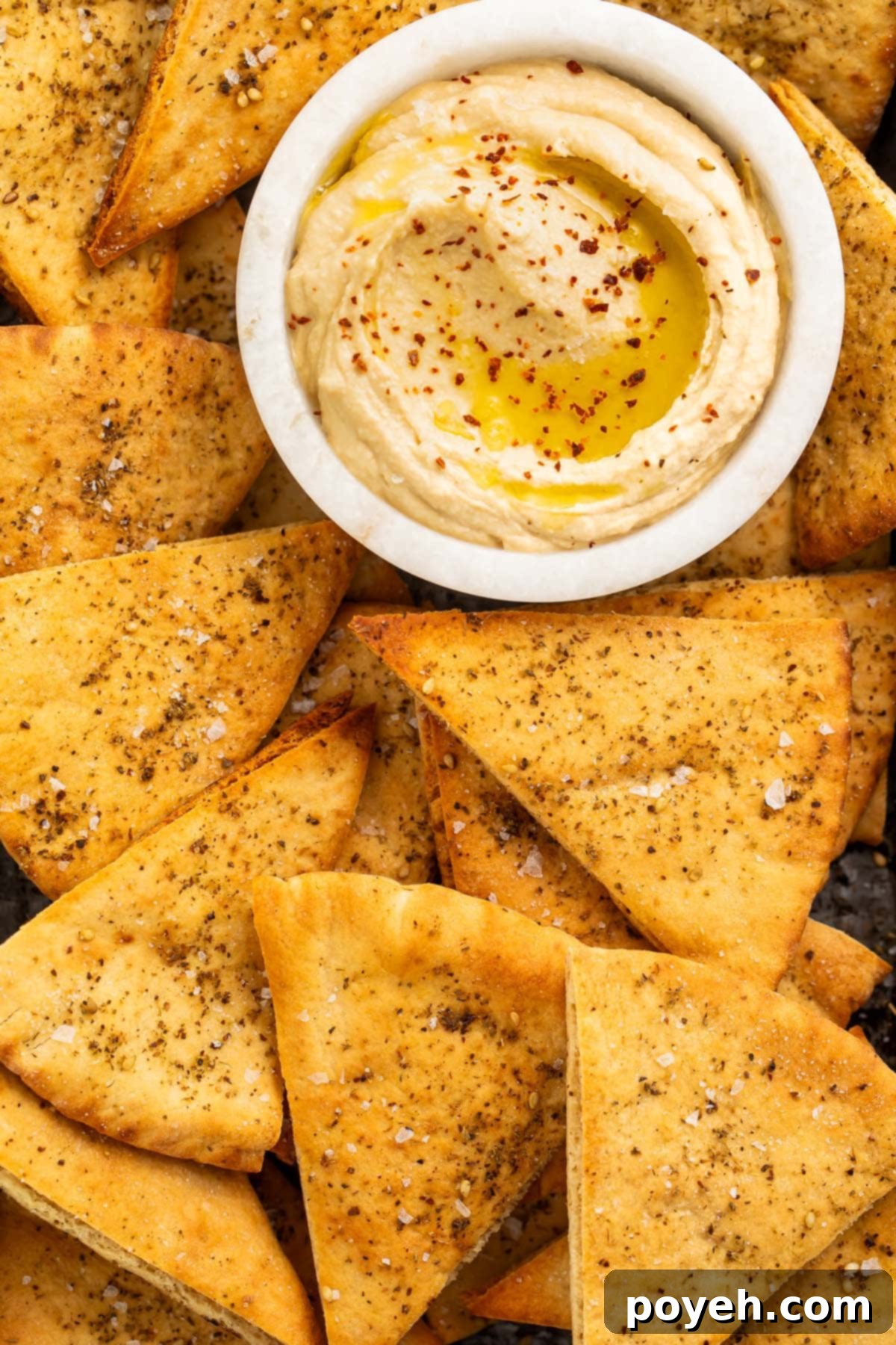 Close-up of golden brown homemade pita chips piled high in a bowl with a small dipping bowl of hummus in the background.