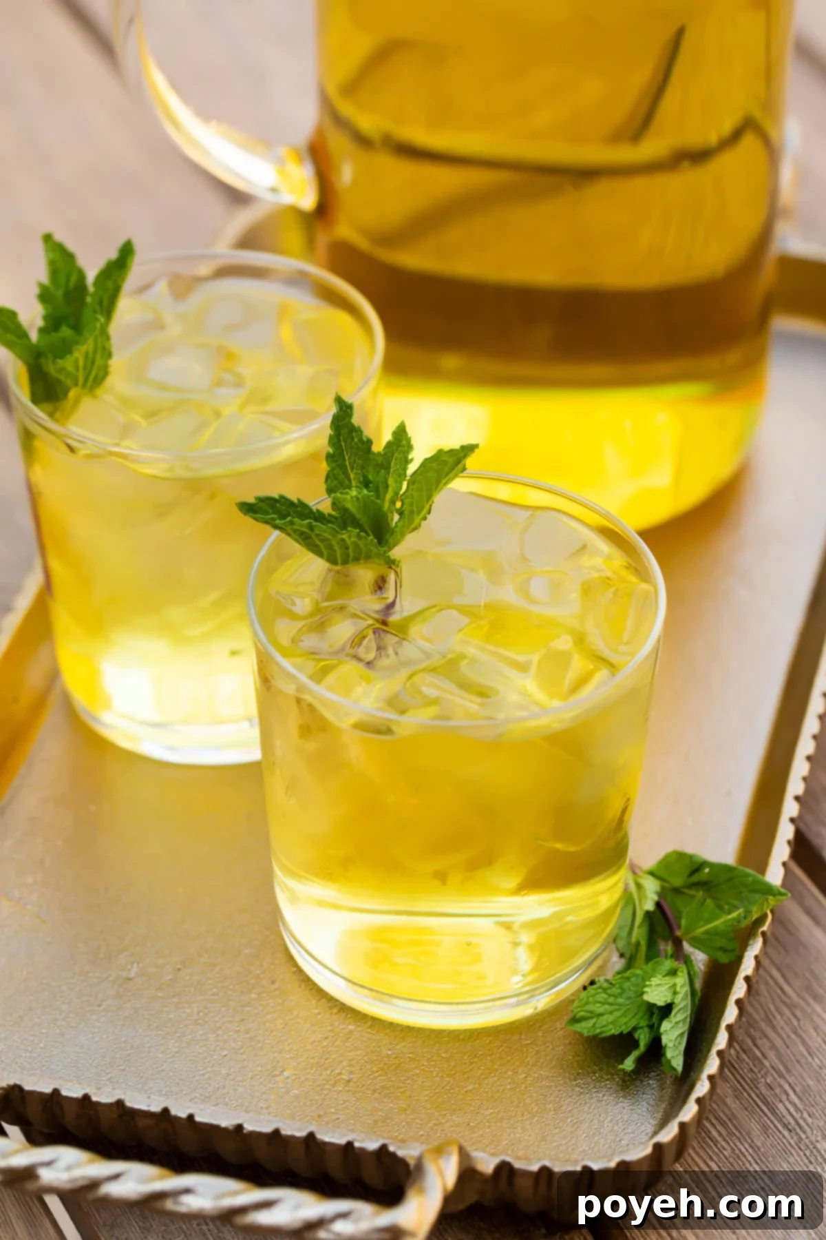 Two elegant glasses of cold brew green tea, garnished with fresh mint and lemon, served alongside a matching pitcher on a rustic wooden table, basking in natural light.