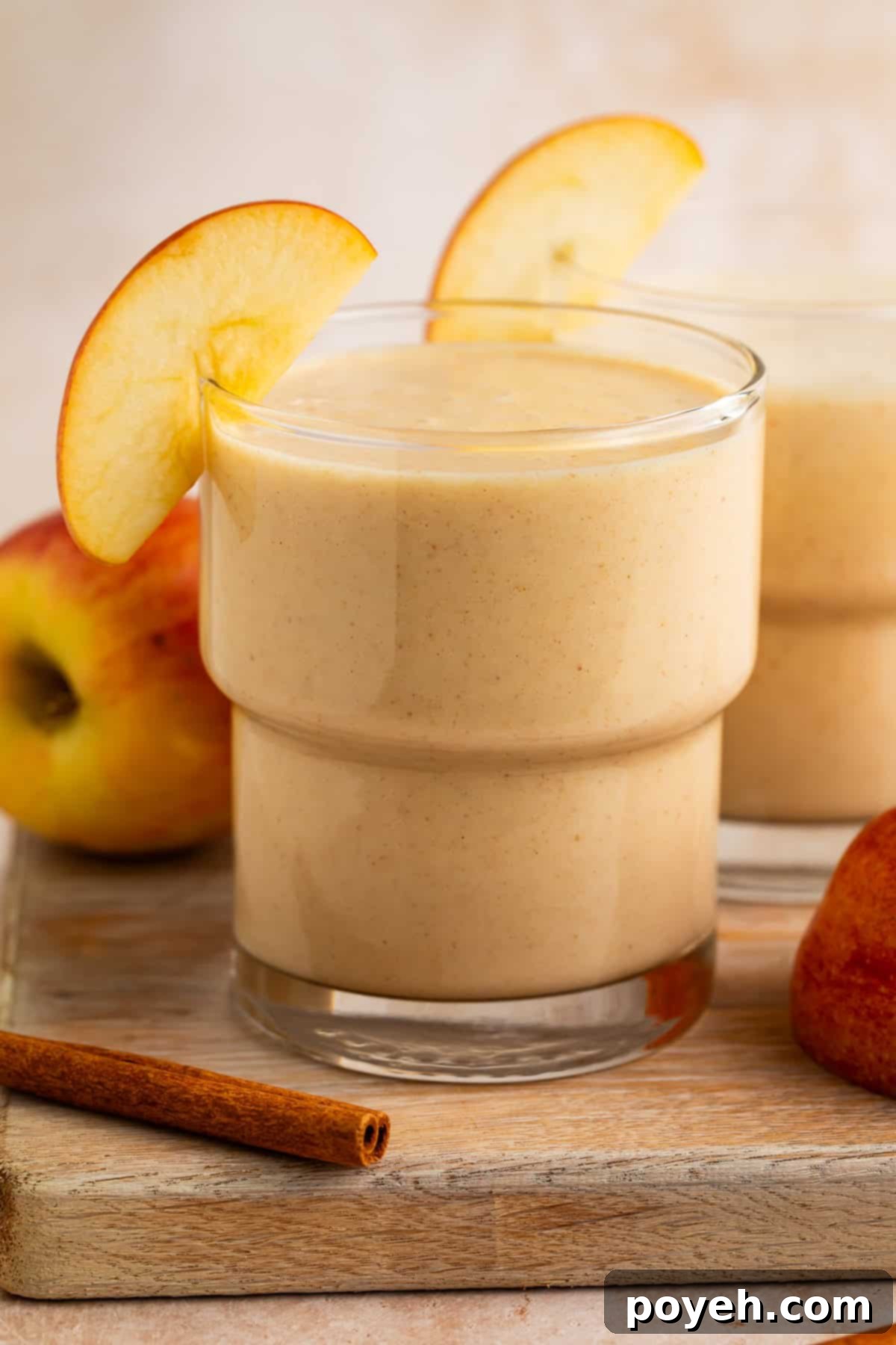 Side on view of an apple smoothie in a glass resting on a wooden board. There are apples and cinnamon sticks next to it.