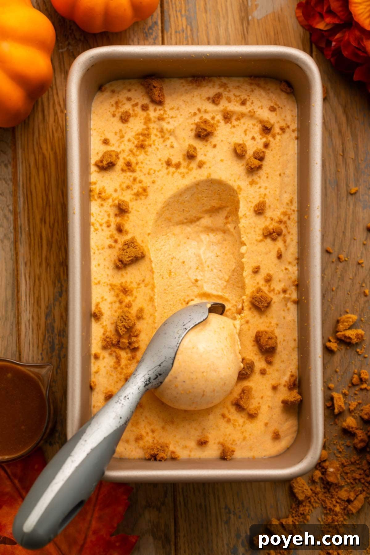 A loaf pan of creamy pumpkin ice cream, with a line of ice cream being scooped from the center of the pan. A silver ice cream scoop holding the scooped ice cream rests on the pan.