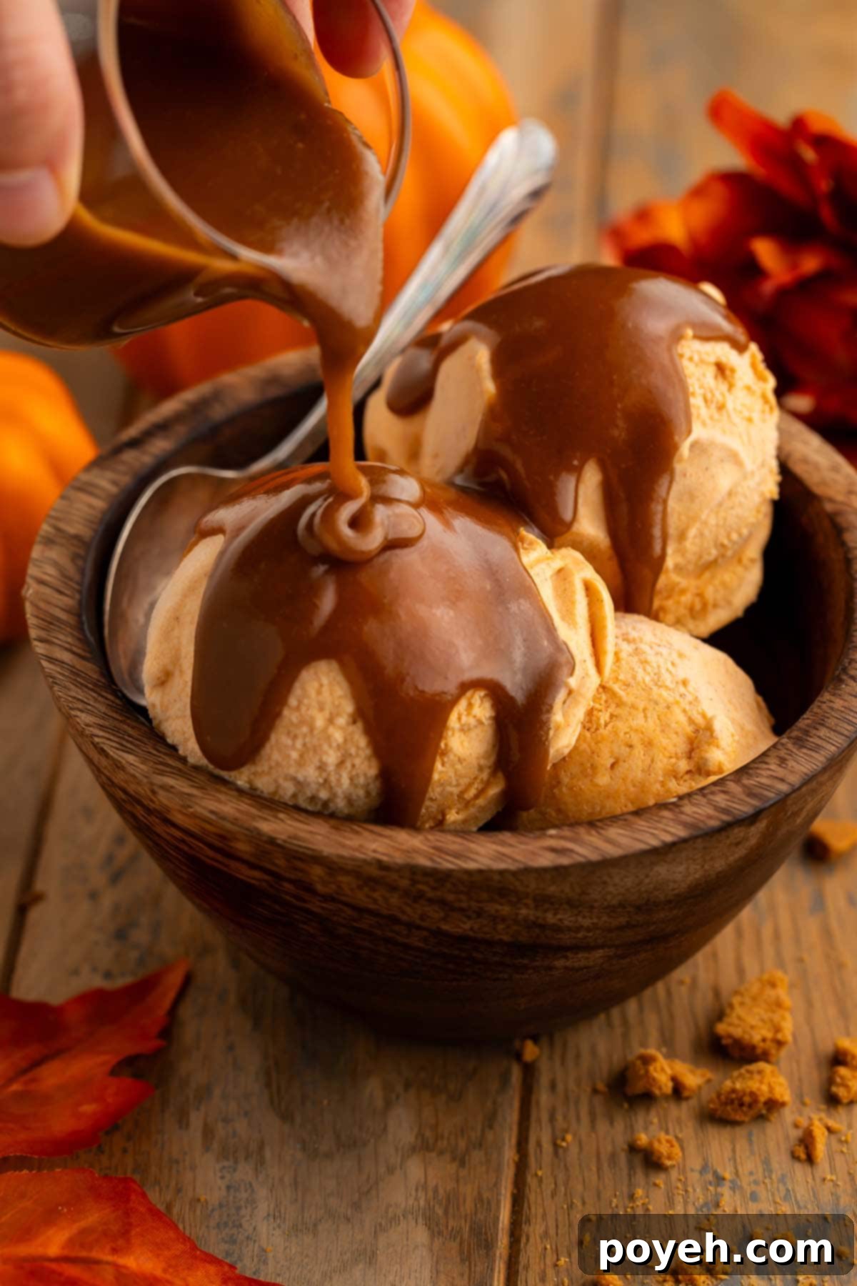 Rich caramel sauce being poured over three scoops of pumpkin ice cream in a turned wood bowl.