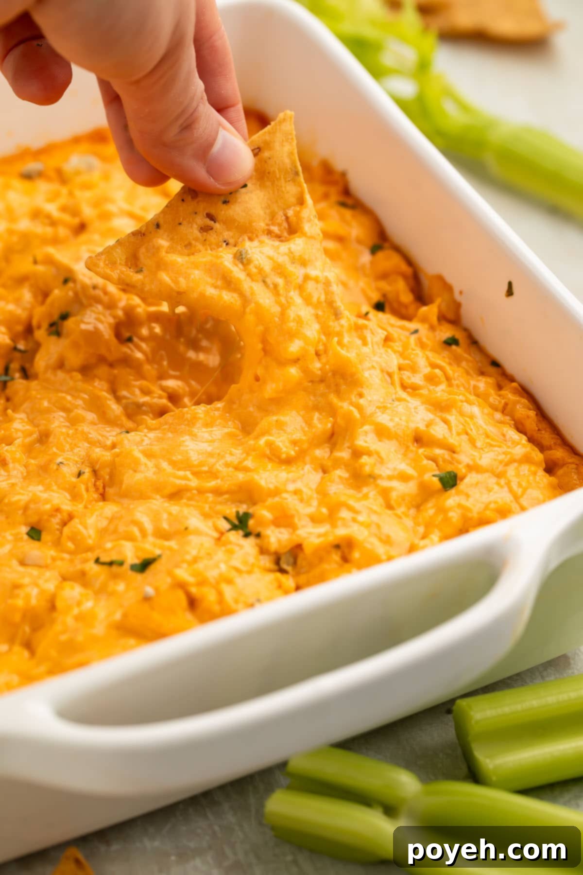 A woman's hand holds a white corn tortilla chip as she uses it to scoop keto buffalo chicken dip out of a white square dish.
