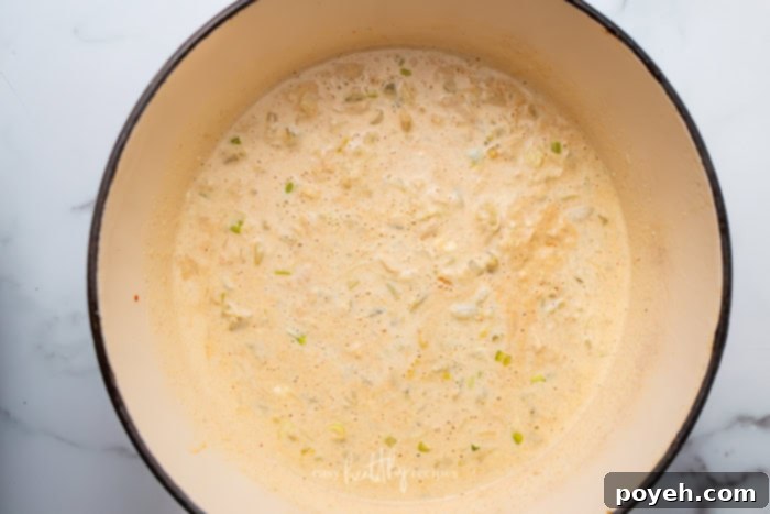 All the creamy chili ingredients, including broth, heavy cream, cream cheese, Sriracha, seasoned chicken, shredded Monterey Jack, and diced green chilis, simmering together in a pot, creating a rich texture.