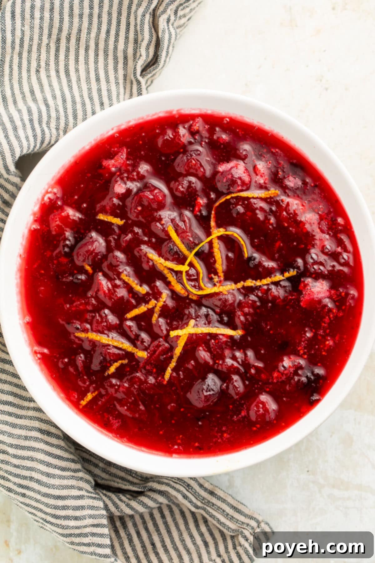 Top-down, overhead view of a white bowl filled with deep red homemade keto cranberry sauce, with whole cranberries throughout, garnished with orange zest.