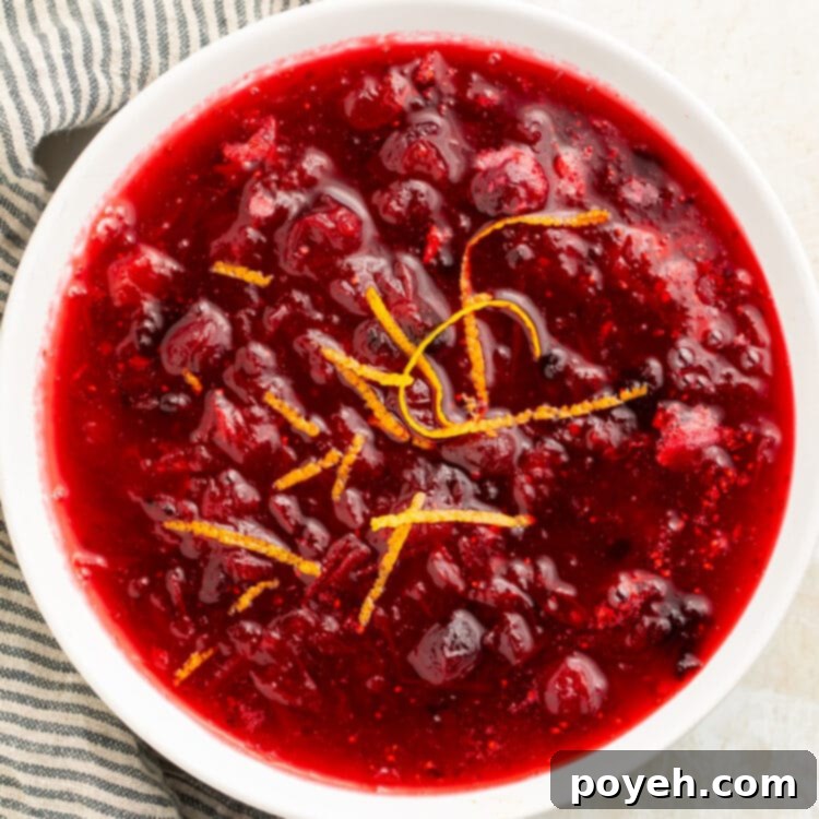 Top-down, overhead view of a white bowl filled with deep red homemade keto cranberry sauce, with whole cranberries throughout, garnished with orange zest.