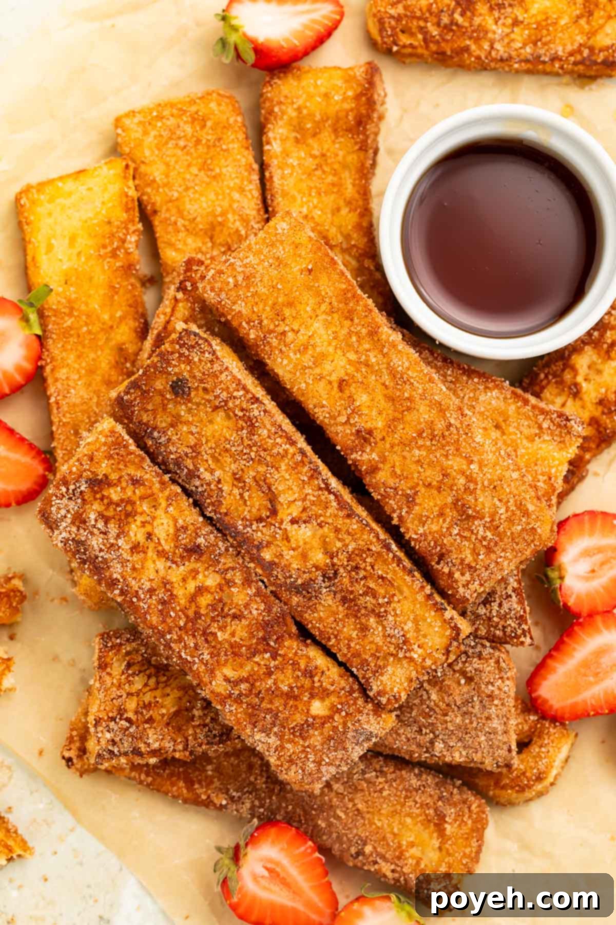 Keto French toast sticks arranged in a pile on a sheet of parchment paper. A ramekin of keto syrup and slices of strawberries sit next to the french toast sticks.