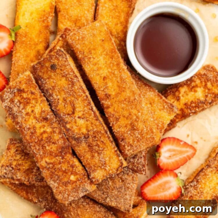 Keto French toast sticks arranged in a pile on a sheet of parchment paper. A ramekin of keto syrup and slices of strawberries sit next to the french toast sticks.