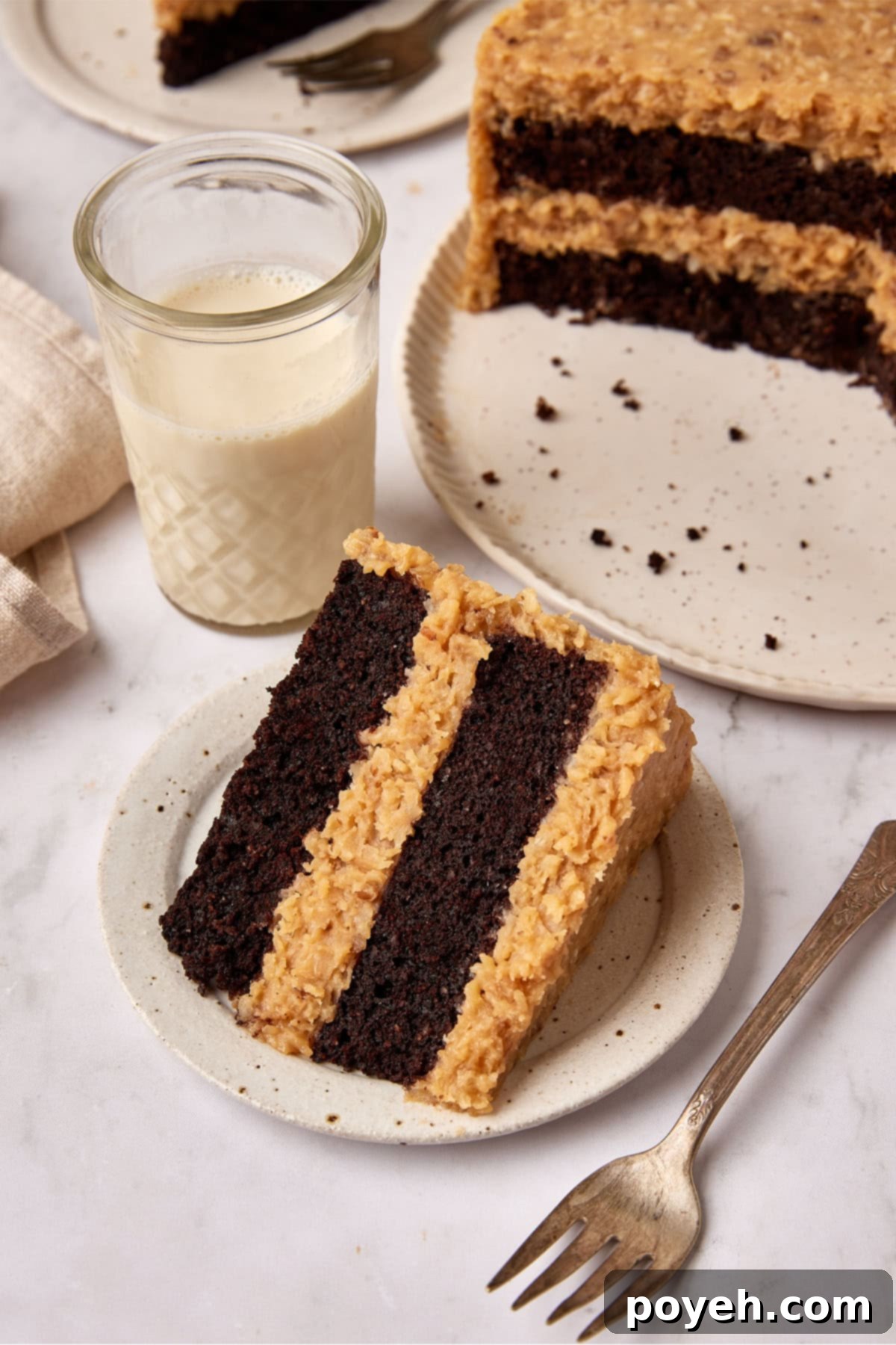 Slice of German Chocolate cake sits on a small white plate next to a fork. A glass of milk is sitting next to it and the cut cake is just visible in the background.
