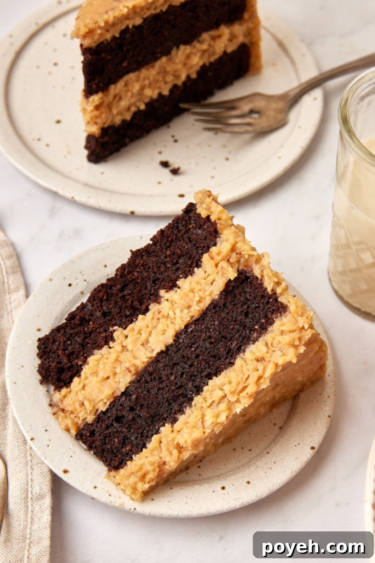 Thick slice of German Chocolate Cake on a white plate, with another slice on a plate with a fork in the background.