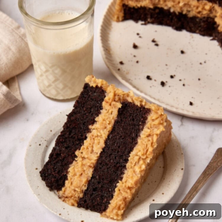 Slice of German Chocolate cake sits on a small white plate next to a fork. A glass of milk is sitting next to it and the cut cake is just visible in the background.
