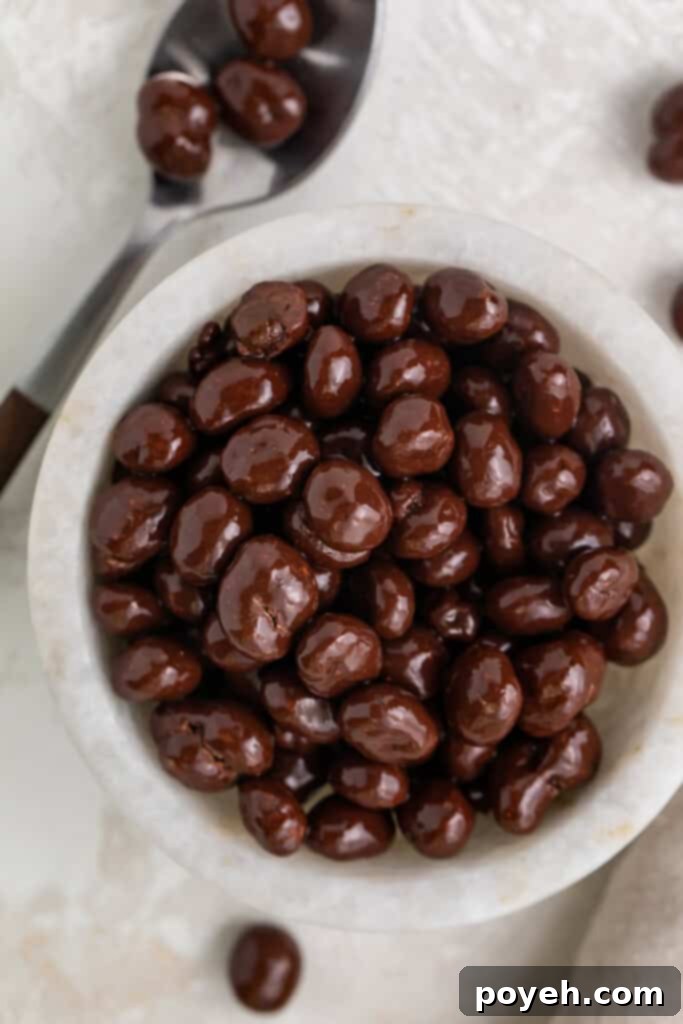 A small white bowl filled with glistening chocolate covered espresso beans, next to a silver spoon, highlighting their rich color and texture.