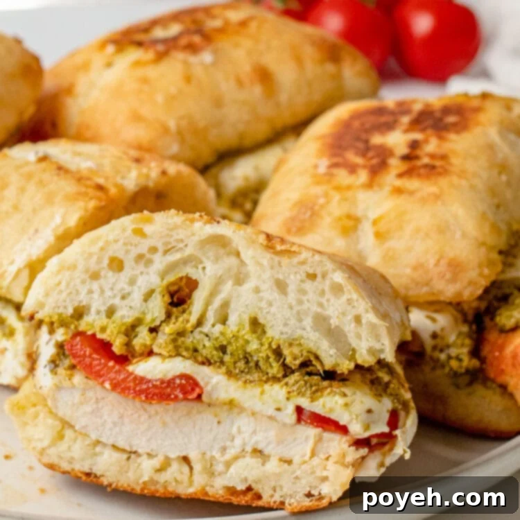 Cross section of a chicken pesto sandwich resting on the lip of a plate, with other sandwiches in the background.